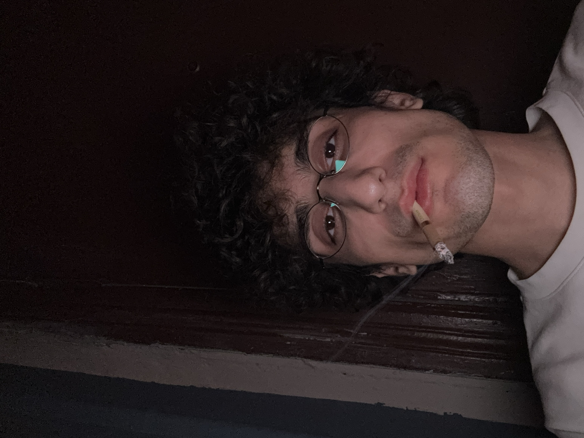 A selfie of a man with curly hair and round glasses, lying down with a lit cigarette in his mouth. The lighting is dim, and the background appears to be a dark wall or ceiling. There is no indication in the image of a location in Moscow, Russia.