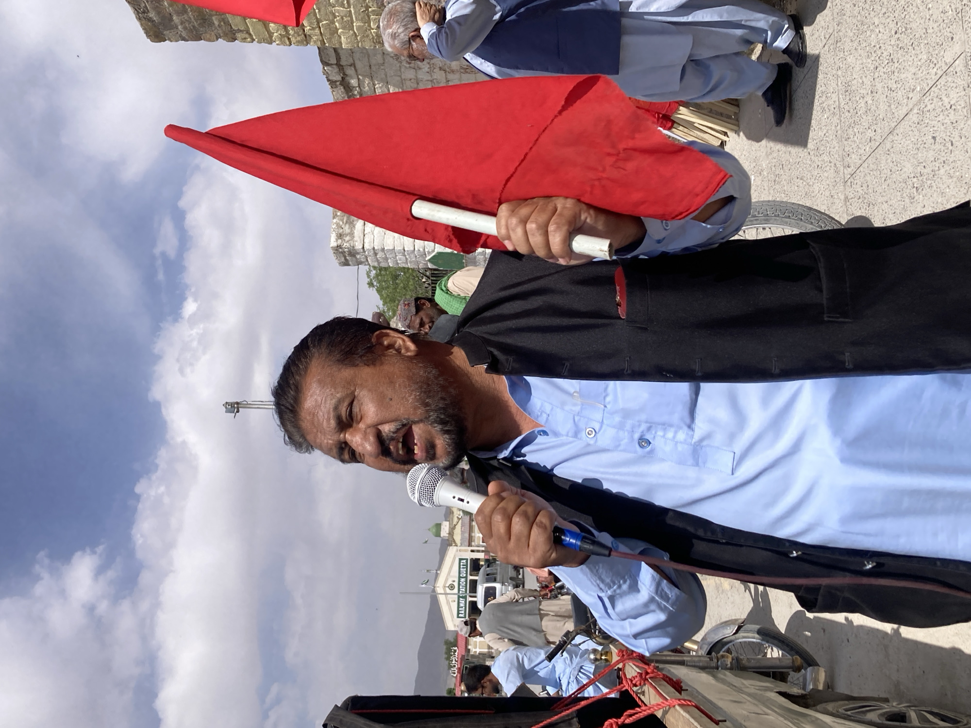 The image captures an outdoor public gathering or protest in Quetta City Tehsil, Pakistan, under a bright, partly cloudy sky. The central subject is a man with a beard, wearing a light blue shalwar kameez and a dark waistcoat. He is actively speaking into a white microphone, his mouth open in mid-sentence, conveying a passionate and engaged demeanor. In his right hand, he holds a red flag attached to a white pole.
Several other individuals are visible in the background and periphery. Another person in the upper right also holds a red flag. Other attendees are dressed in traditional South Asian attire, such as shalwar kameez.
The event takes place on paved ground, with a partial view of what appears to be a vehicle or cart, featuring ropes and a tire, in the lower right foreground. In the mid-ground, a prominent white building with a green dome serves as a key location marker. A sign on this building clearly reads "RAILWAY STATION QUETTA" in English, confirming the specific city. Below the English text, additional script in Urdu or Arabic is visible, reading "لا إله إلا الله" (La ilaha illallah), which translates to "There is no god but Allah." Distant mountains frame the horizon under the blue sky, indicating the geographical setting of Quetta. The overall scene suggests a public address, rally, or demonstration.