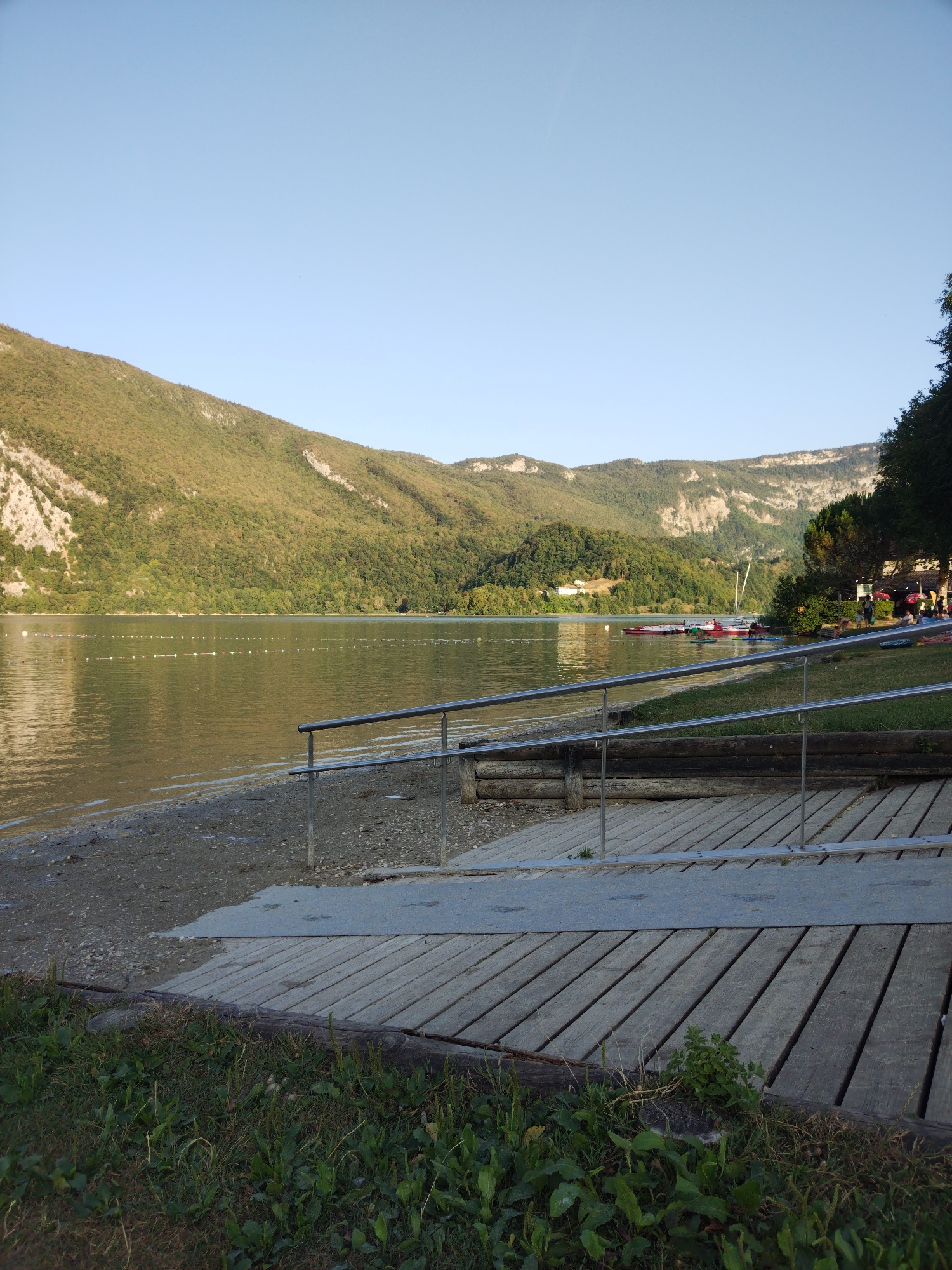 A wide shot captures a serene lake scene in Novalaise, France, on a clear, bright day. The calm, reflective water dominates the left side of the frame, with a gentle shoreline of pebbles and sand visible in the foreground. A wooden ramp with metal railings leads down to the water's edge, suggesting accessibility. Beyond the lake, a verdant, tree-covered mountain range rises majestically, its slopes catching the sunlight. In the distance, several small boats are moored or in motion on the lake, and a few people can be seen on the grassy bank to the right, near a building and red-umbrella-covered tables, indicating a recreational area or cafe. The sky above is a clear, pale blue, with no clouds visible.