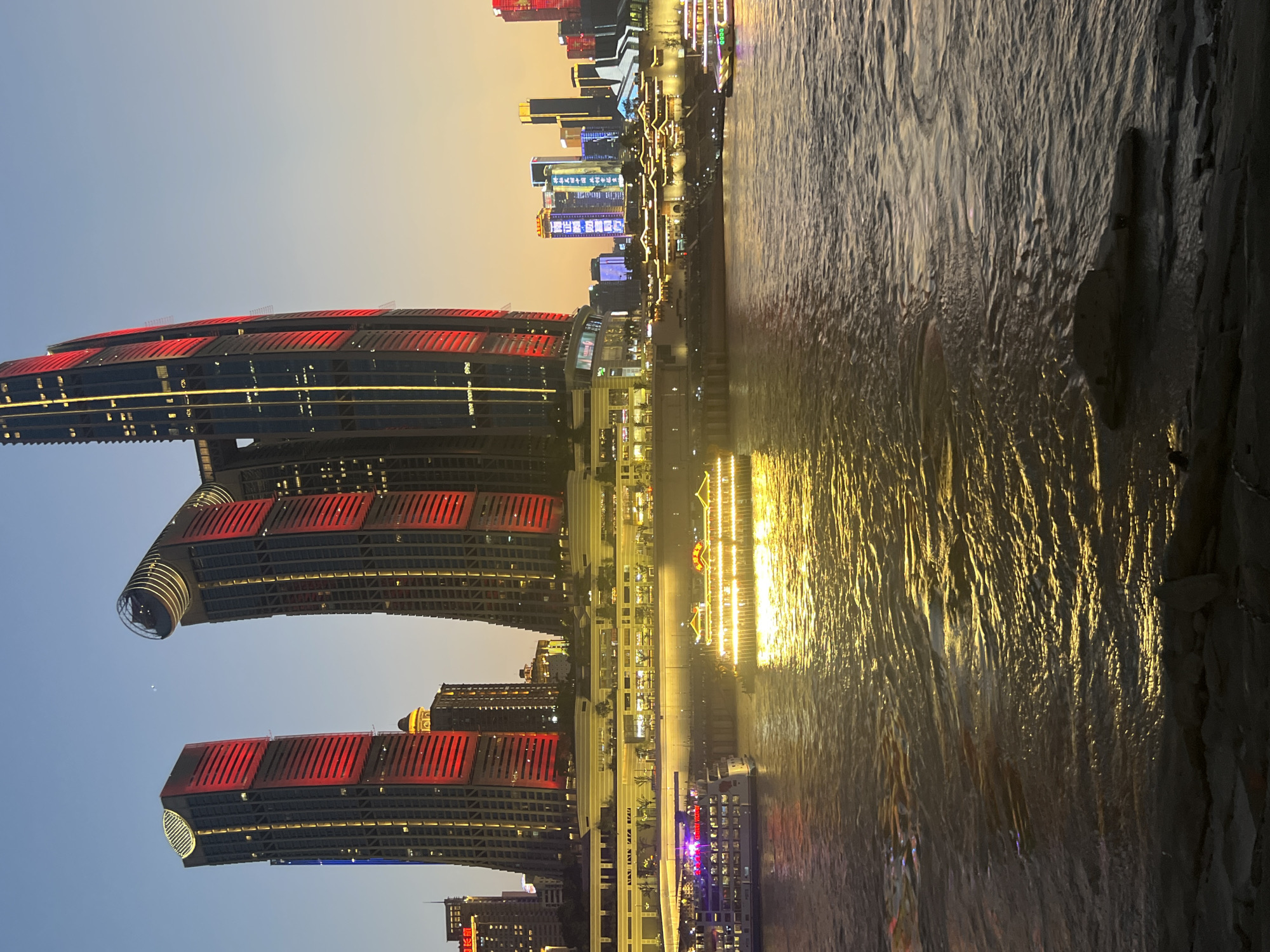The image depicts a waterfront scene in Nan'an District, Chongqing, China, during twilight. Two prominent skyscrapers, illuminated with red and yellow lights, dominate the skyline. The left skyscraper features a rectangular design with red lights on its upper section, while the right skyscraper has a more curved, cylindrical top also lit with red and yellow. Below these structures, a series of buildings with traditional Chinese architectural elements are visible, illuminated with warm yellow lights, suggesting a commercial or entertainment area.
A river flows in the foreground, reflecting the lights from the buildings and two boats. The boat on the left is larger and appears to be a passenger vessel, displaying purple and white lights. To its right, a smaller, ornate boat, possibly a restaurant or tour boat, is brightly lit with golden lights, casting a strong reflection on the water. The water's surface is rippled, and the reflections create a shimmering effect. The sky above transitions from a deep blue at the top to a warm orange hue near the horizon, indicating either sunset or sunrise. The riverbank in the foreground is rocky and dark. The overall impression is one of a vibrant urban landscape at dusk.