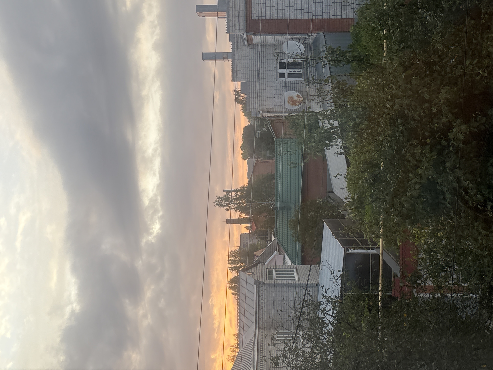 The image depicts a view of a residential area in Georgiyevsk, Russia, during what appears to be sunset or sunrise. The sky is filled with clouds, with hues of orange and yellow visible near the horizon, indicating twilight. In the foreground, dense green foliage of trees obscures parts of the scene. Several houses with pitched roofs, some covered in metal sheeting and others with green shingles, are visible. One building on the left is constructed of light-colored brick with grey metal roofing. Another building to the right is made of white brick and features two satellite dishes mounted on its facade. Chimneys or ventilation pipes extend vertically from the roof of the white brick building. In the background, more residential structures and trees are visible, along with a tall, industrial-looking chimney or tower. Electrical wires are strung horizontally across the upper portion of the image. No people or vehicles are discernible in the scene.