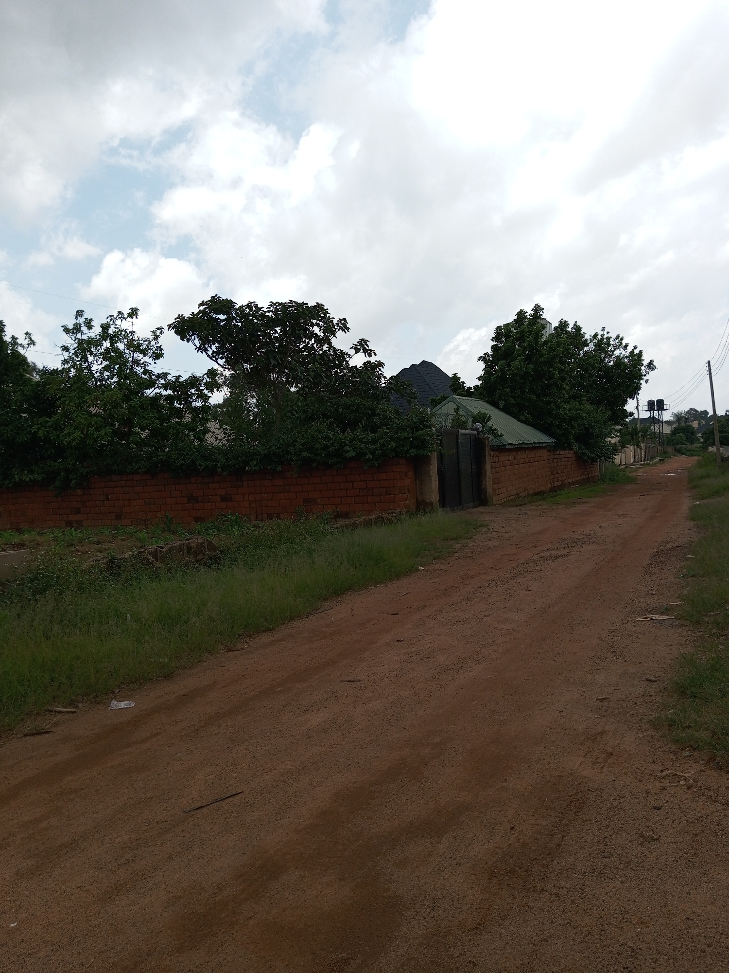 The image depicts a dirt road in Gura Top, Nigeria, with a brick wall on the left side lined with trees and vegetation. Beyond the wall, a house with a green roof and black gate is visible, partially obscured by trees. In the distance, several other structures and trees are scattered across the landscape, with power lines running overhead. The sky is cloudy, with patches of blue visible. The road appears to be a rural or semi-urban path, possibly leading into a residential area. The overall scene suggests a warm, possibly humid climate.