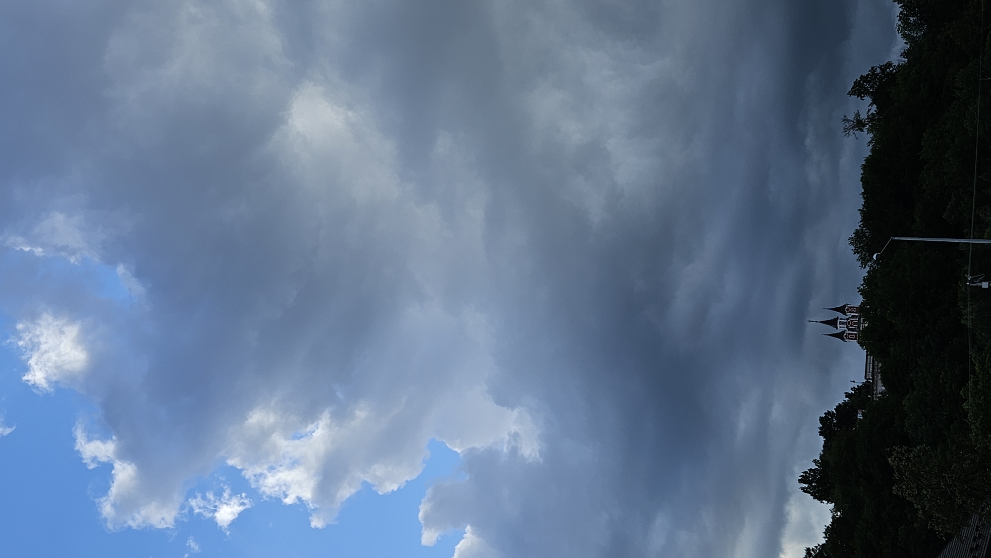 This media file depicts a dramatic sky with ominous, dark clouds dominating the frame. The clouds are a mix of grays and blues, suggesting an approaching storm or overcast weather. In the lower right portion of the image, partially obscured by dark green trees, is a building with distinctive spires, hinting at a church or a similar architectural structure. A lamppost is also visible to the right of the building, suggesting an urban or suburban setting. The scene appears to be outdoors, with the sky occupying the majority of the composition. The time of day is difficult to ascertain precisely due to the heavy cloud cover, but the lighting suggests it could be late afternoon or early evening. There are no people or discernible activities taking place. The overall mood conveyed is one of impending weather change, with a sense of drama and perhaps a touch of foreboding due to the dark clouds. The location context provided places this scene in Prešov, Slovakia.