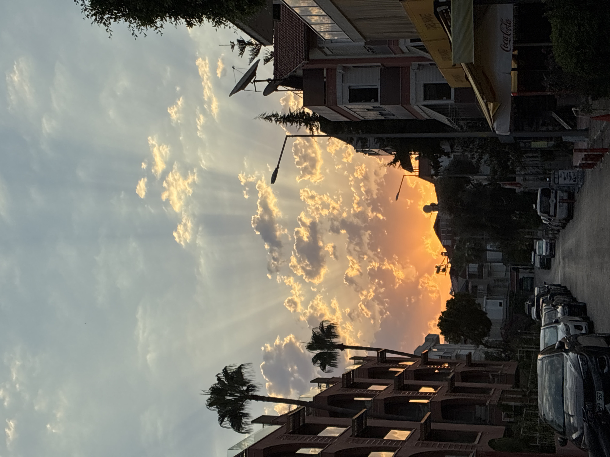 The image captures a street scene in Alanya, Turkey, at sunset. The sky is dominated by dramatic crepuscular rays beaming through scattered clouds, illuminated by the bright orange glow of the setting sun. On the left, a multi-story building with arched windows and balconies is visible, flanked by palm trees. On the right, other buildings, some with satellite dishes on their roofs, line the street. A street lamp stands prominently. Several cars are parked along the asphalt road, and a cafe with a yellow awning displaying the Coca-Cola logo is situated on the right side of the street. The overall atmosphere is serene and picturesque, indicative of a Mediterranean evening.