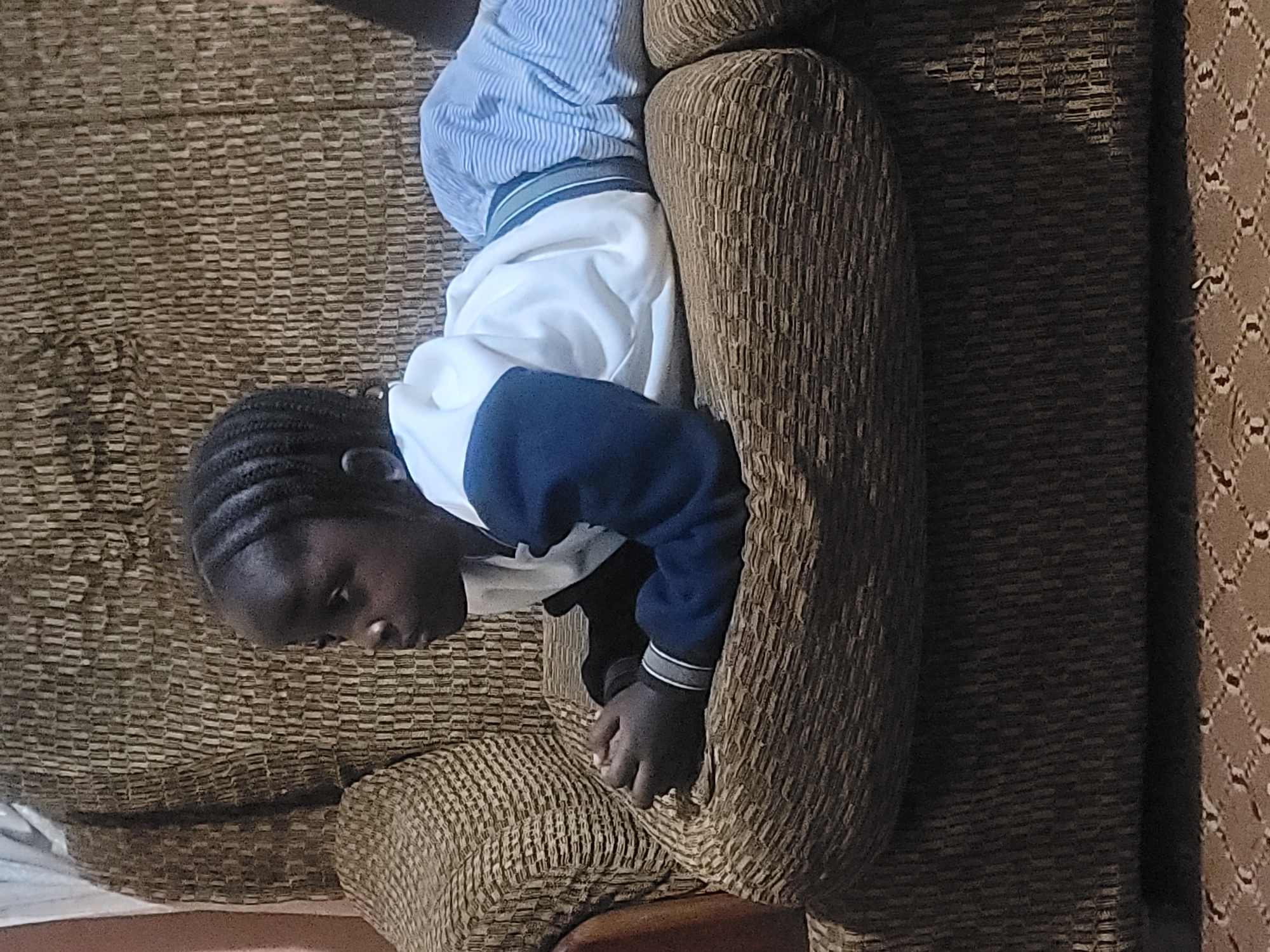 A young Black child with braided hair lies prone on a textured brown sofa. The child wears a white hooded sweatshirt with blue sleeves and blue and white striped pants. The child's hands are clasped in front of their chest. The sofa has a woven pattern. The background is largely obscured by the sofa and a portion of a light-colored wall. The lighting is dim, casting shadows. The image is taken from a medium distance, focusing on the child and the immediate area of the sofa. The location is an indoor domestic setting.
