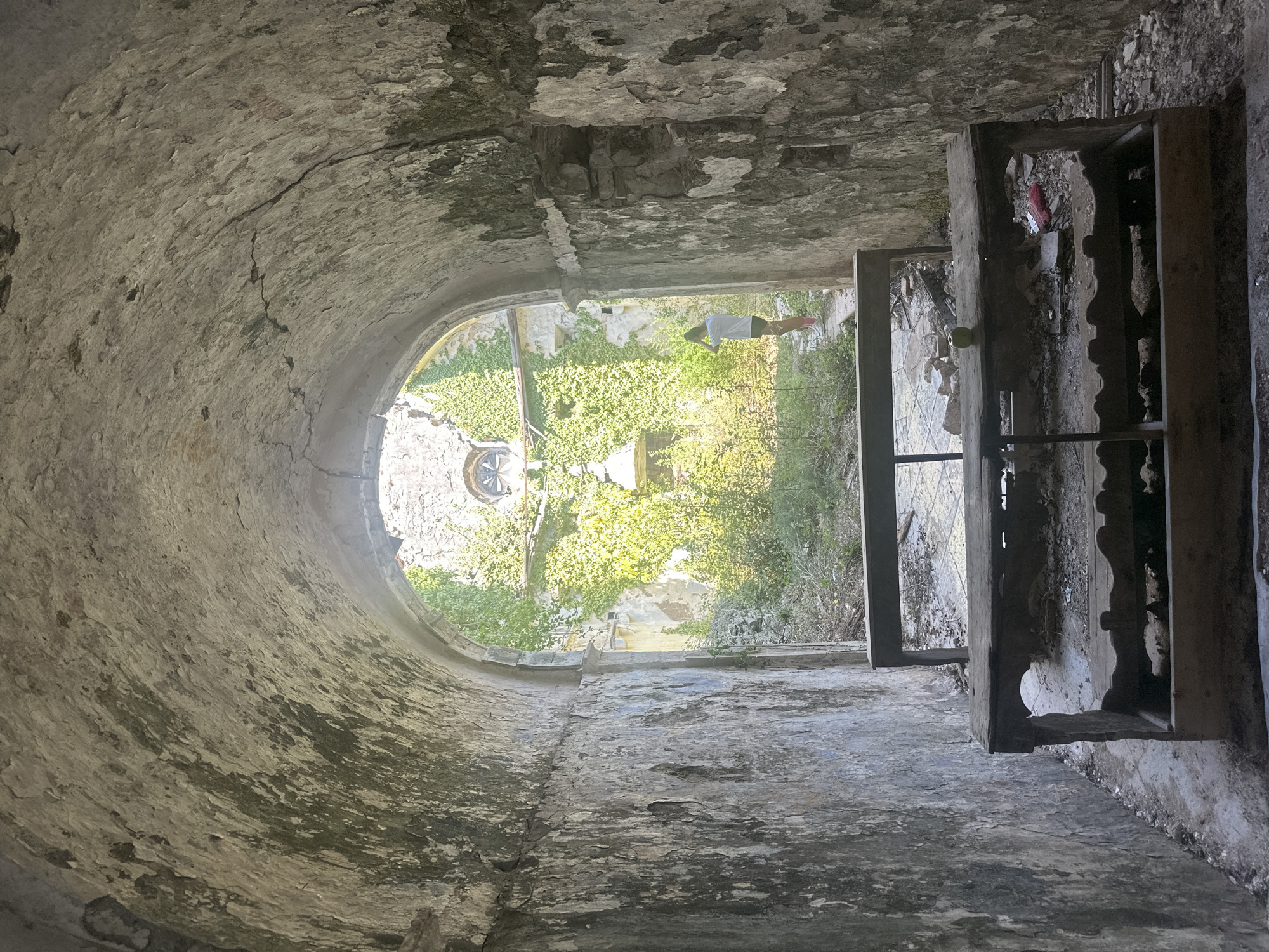 The image depicts the interior of a stone tunnel or arched passageway, looking out towards an overgrown courtyard. The walls of the tunnel are rough and weathered, showing signs of age and decay with moss and discoloration. In the foreground, a dark, wooden bench with two levels of shelving is positioned centrally. A single green apple rests on the top surface of the bench. To the right of the bench, a crumpled red object, possibly a wrapper, is visible among debris.
Beyond the tunnel's archway, a sunlit courtyard is filled with dense green foliage and vegetation, suggesting neglect or abandonment. Ivy covers the stone walls of a building in the background, where a circular window with intricate stonework is visible. Standing in the courtyard, a young person wearing a white t-shirt, dark shorts, and pink socks is facing away from the camera, with their hands raised to their head. The ground in the courtyard appears to be paved with stone tiles, interspersed with rubble and vegetation. The overall atmosphere is one of ruin and nature reclaiming man-made structures. The location is identified as the abandoned seminary in Prčanj, Montenegro.