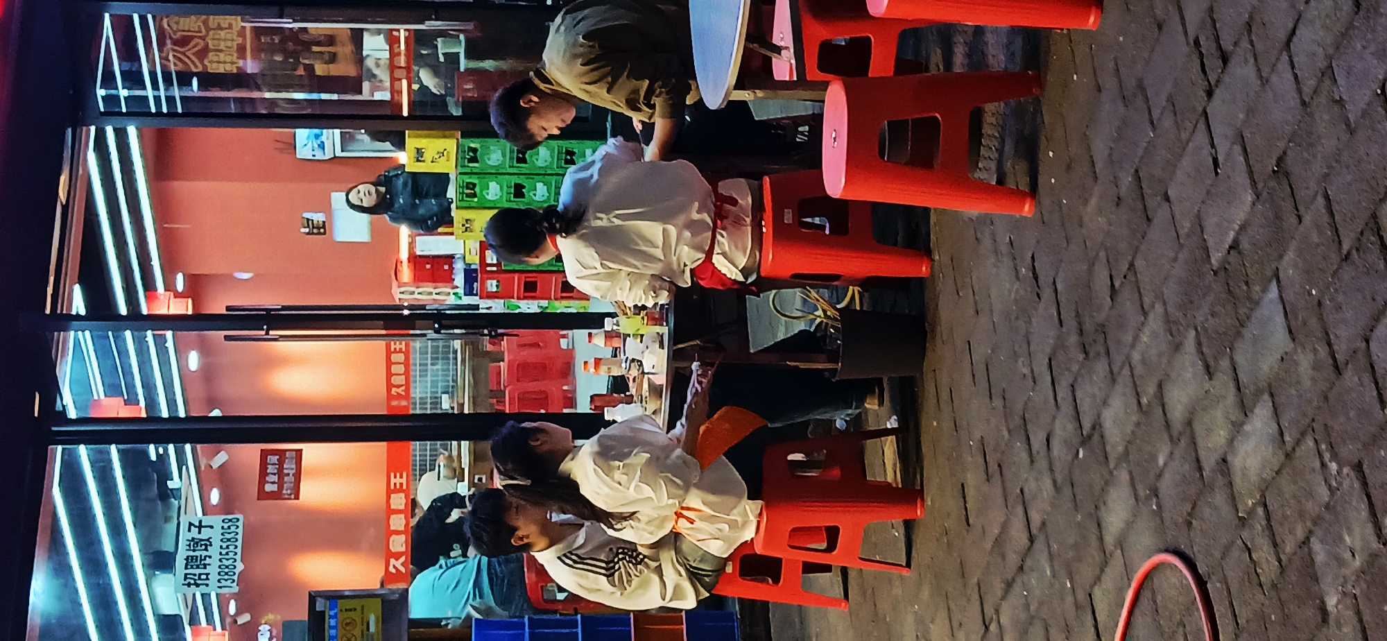 The scene depicts a casual dining setting in Jiulongpo District, China, outside a restaurant at night. Three couples are seated around outdoor tables, likely dining, each person on a red plastic stool. The foreground is the textured pavement. The restaurant's interior, seen through the glass front, has red walls, a sign with Chinese characters, and stacks of colorful crates. A person in a black jacket is visible in the reflection inside. The storefront has illuminated white vertical lines and a recruitment sign with a phone number. The lighting is a mix of artificial lights from the restaurant and the outdoor environment.