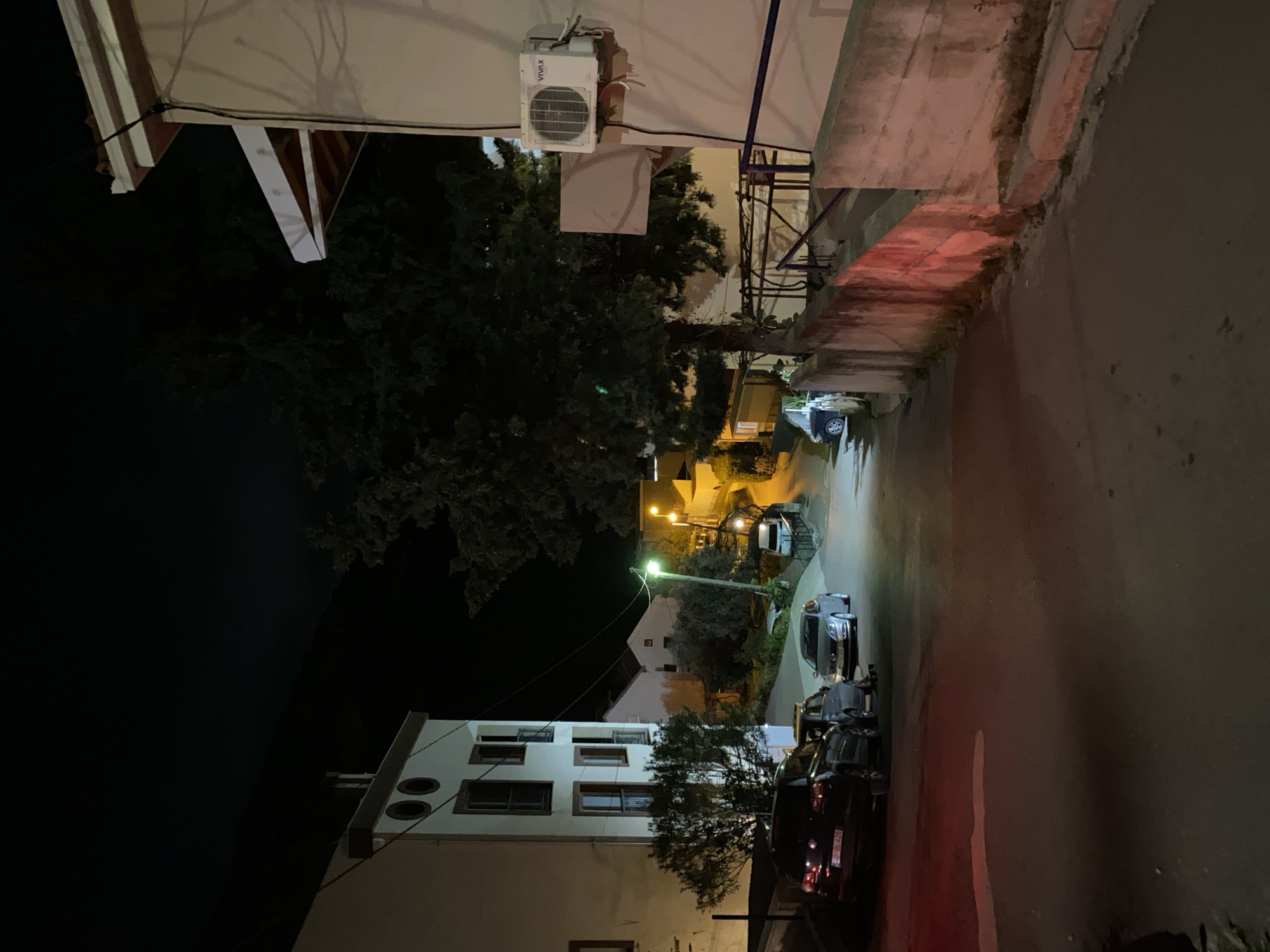 The image shows a narrow street in Budva, Montenegro at night. The street is flanked by buildings on both sides. On the left side of the street is a light-colored building with multiple windows and an air conditioning unit affixed to its exterior wall. A tree partially obscures the view of this building. On the right side of the street is a taller, weathered concrete structure. Cars are parked along the street, with some parked closer to the viewer. Streetlights illuminate the scene, with one particularly bright light located further down the street. The overall lighting is dim, with shadows cast by the buildings and trees. The street appears to be relatively quiet.