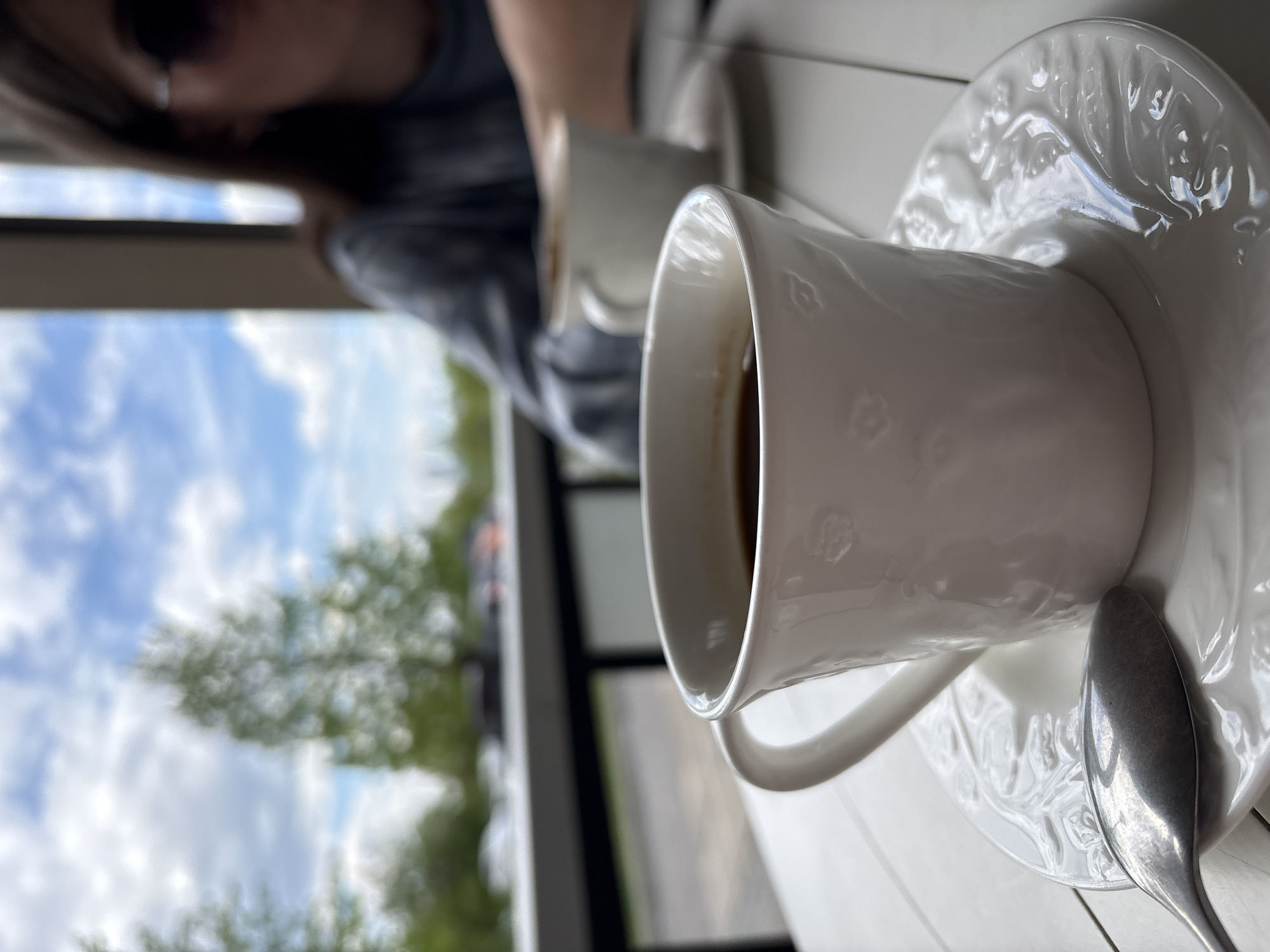 The image captures a moment in a cafe or restaurant, possibly in Krasnoyarsk, Russia, indicated by the partially visible cityscape or landscape through the window. In the foreground, a white porcelain coffee cup sits on a matching ornate saucer, accompanied by a silver spoon. The cup appears to contain a dark liquid, likely coffee or tea. Another cup and saucer are partially visible behind the first, suggesting a setting where people are enjoying beverages. A person, only partially in the frame and wearing sunglasses, is positioned near the window, possibly enjoying the view. The overall scene suggests a relaxed, social atmosphere within an indoor establishment.