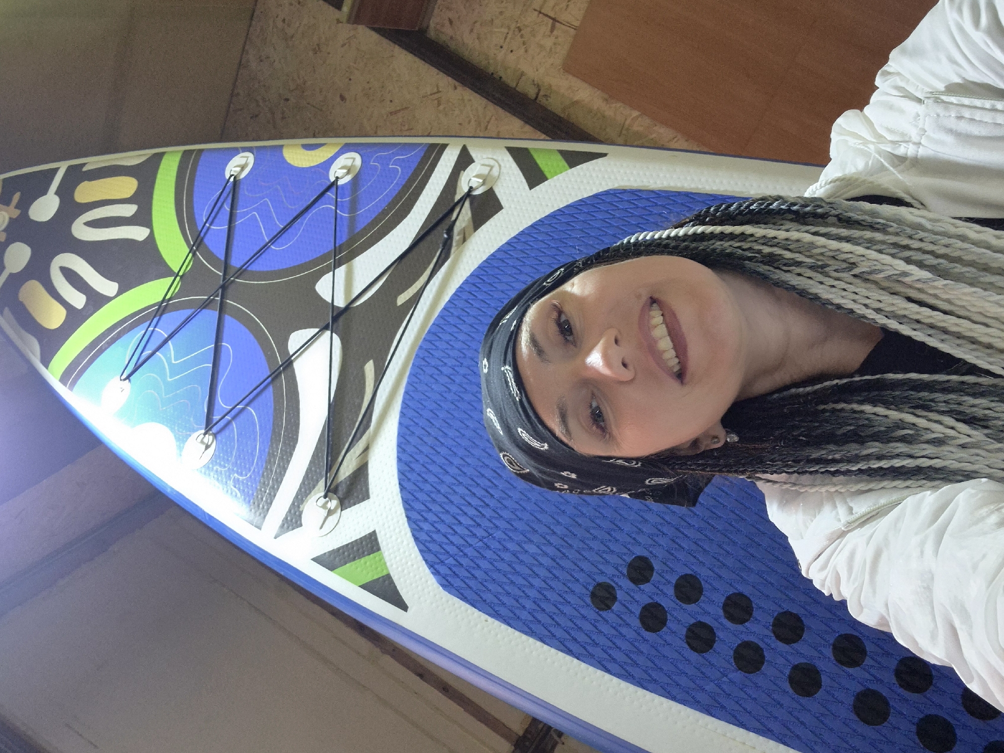A woman with long, braided hair, black bandana, and white jacket smiles at the camera, standing in front of a colorful paddleboard. The paddleboard has a blue and white base with black dots and a patterned top with green, blue, and white designs. The background appears to be an indoor space, possibly a garage or storage area, with wooden walls and possibly some stored items. The scene does not provide any specific indication of its location within the city of Chaikovsky, Russia.