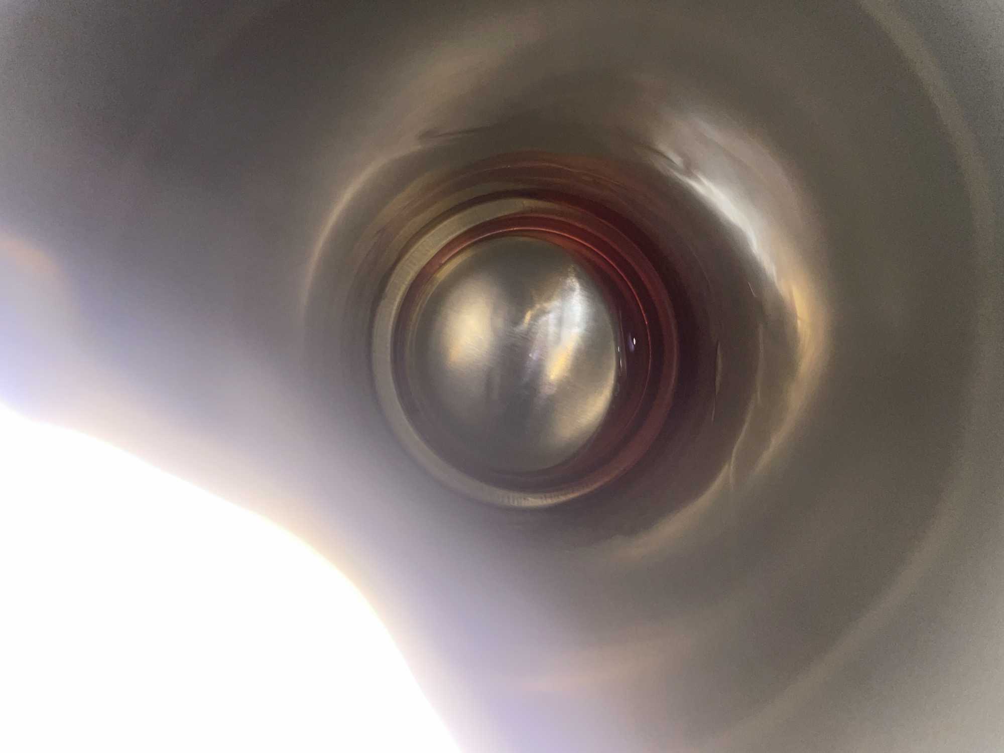 The image is a close-up, distorted view from inside a metallic object. The metallic surface is curved and reflects light, creating a glossy, almost liquid appearance. The central focus is a circular opening, within which concentric rings of red and silver are visible. Beyond this opening, a bright, hazy light emanates, suggesting an external light source or an opening to a brighter space. The overall impression is one of looking into a tunnel or a hollow cylindrical object, with the camera positioned at one end and looking towards the other. The perspective is disorienting due to the curved reflections and the close proximity to the surface. No people or specific environmental features are identifiable within the frame, only the abstract interplay of light and form on the metallic interior.