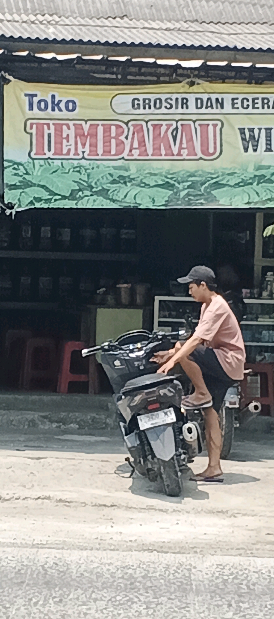 A person wearing a light pink t-shirt, dark shorts, a dark baseball cap, and flip-flops is sitting on a parked scooter. The scooter is dark gray and has a license plate that reads "B 2000 XY". The person's right leg is bent, with their foot resting on the scooter's body. Their hands are on the handlebars. The scooter is parked on a sandy, slightly rough surface in front of a store with a large green and yellow banner. The banner has text that appears to say "Toko Grosir Dan Ecera Tembakau Wi". Behind the scooter, there are shelves with various items visible inside the store. Red chairs are also visible through the store's opening. The overall scene suggests a street-side shop in the city of Sumberwudi, Indonesia.