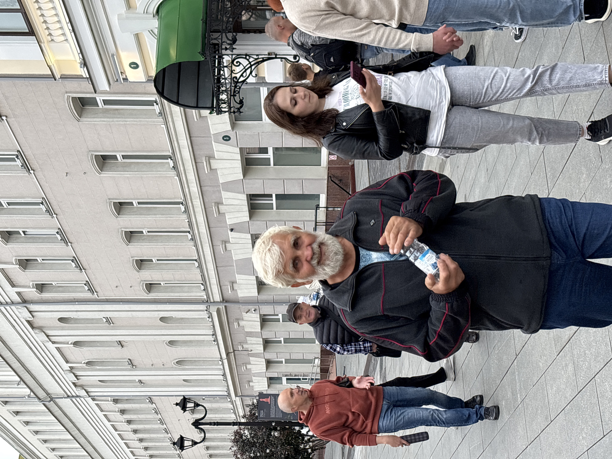 This image captures a street scene in Nizhny Novgorod, Russia. Several people are visible, some in the foreground and others further back. In the immediate foreground, a man with a grey beard is looking towards the camera, holding a bottle of water. He is wearing a dark jacket with red trim and blue jeans. Behind him, another man in a red hoodie and jeans is looking to the left. Further back, a woman with long brown hair is holding a smartphone and wearing a black leather jacket over a white t-shirt and grey jeans. Other individuals are partially visible in the background, suggesting a public gathering or a casual stroll.
The setting appears to be a pedestrian area with a building facade featuring numerous windows and architectural details. A green awning is visible on the building above the woman with the phone. Two streetlights are also present, suggesting it might be daytime, although the lighting is somewhat overcast, perhaps indicating a cloudy day. The ground is paved with grey tiles. There is no explicit text visible on the people's clothing, but the water bottle has some text that is too small to read. The overall mood seems relaxed and everyday.