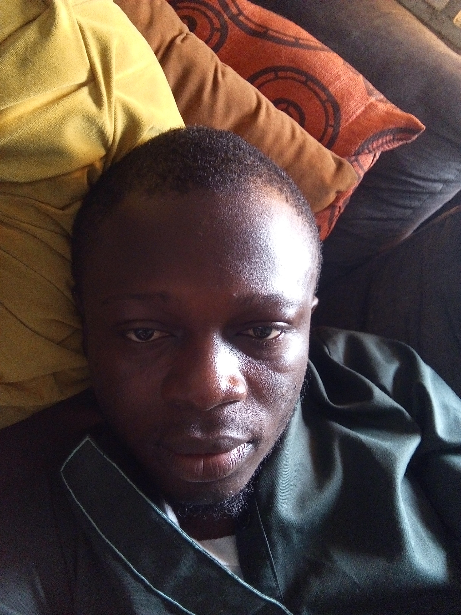 The image displays a close-up, high-angle portrait of an adult male with a dark complexion, positioned horizontally as if lying down. His face is centered and directed towards the camera, with a neutral, relaxed expression and semi-lidded eyes. He has short, dark, closely cropped hair and a faint goatee on his chin. Significant highlights are visible on his forehead, nose, and right cheekbone (viewer's left), suggesting a light source from the upper left.
He is wearing a dark, possibly forest green, collared garment with a visible lighter-colored piping or trim along the lapel and a white or light grey undergarment at the neckline. The fabric of his outer garment shows soft folds and creases.
The background comprises several cushions or pillows in an indoor setting. To the left of the man's head is a bright yellow, heavily folded cushion. Further back and to the right, a plain brown cushion is visible, alongside an orange cushion featuring a repeating dark, circular, concentric pattern. The furthest background element appears to be a darker, possibly grey or black, upholstered surface, indicative of a sofa or bed.