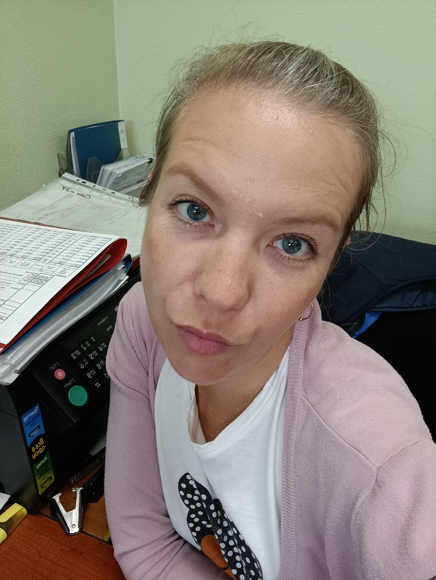 The image shows a close-up, head-and-shoulders selfie of a white woman with blonde hair pulled back, blue eyes, and fair skin. She is wearing a white t-shirt with a black and white polka dot pattern and a light pink cardigan. She is looking directly at the camera with a slightly puckered expression.
The background suggests an office environment. To the left, there is a stack of papers and folders, a black office machine with buttons and labels (some in Cyrillic script), and a stapler. Behind the woman, a dark blue jacket or piece of clothing is visible. The wall in the background is a pale green color.
The location is identified as the city of Chaikovsky, Russia, suggesting this office is within that city. The overall scene is a casual office selfie, likely taken at a desk.