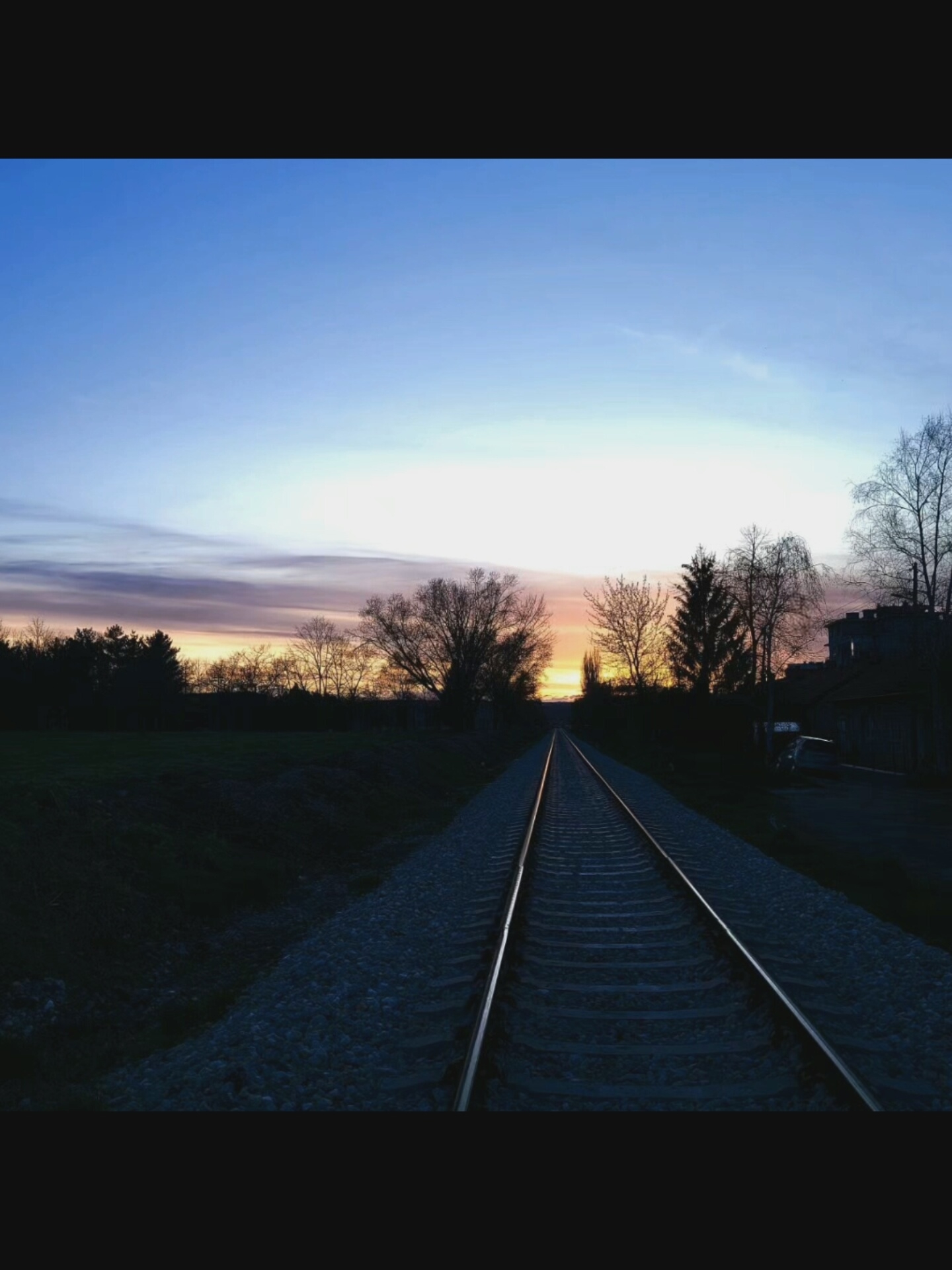 An outdoor, low-angle, eye-level shot captures a vanishing point perspective of railroad tracks stretching into the distance. The sky above is a gradient of twilight colors, transitioning from a clear, deep blue at the top to soft hues of pink and orange near the horizon, indicating either dawn or dusk. Silhouetted trees and buildings line the right side of the tracks, with a dark wooded area visible on the left. A faint light from a structure on the right suggests artificial illumination. The scene appears to be in Bielawska Wieś, Poland.