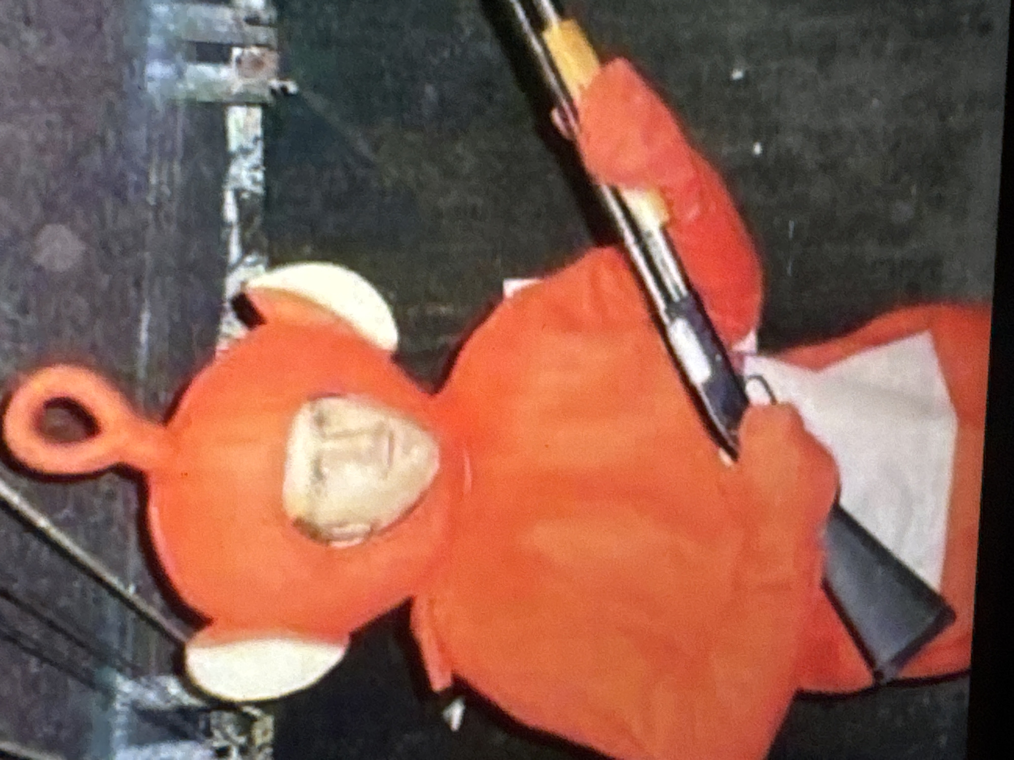 A man in an orange Teletubby costume is holding a shotgun. The man is seen from the chest up, with the orange costume covering his entire body except for his face. His face is visible through an opening in the costume's headpiece. He is holding the shotgun with both hands, pointing it downwards and slightly to his right. The background appears to be an indoor setting with a dark, textured wall. There is no indication of the location being in Kuta Selatan, Indonesia.