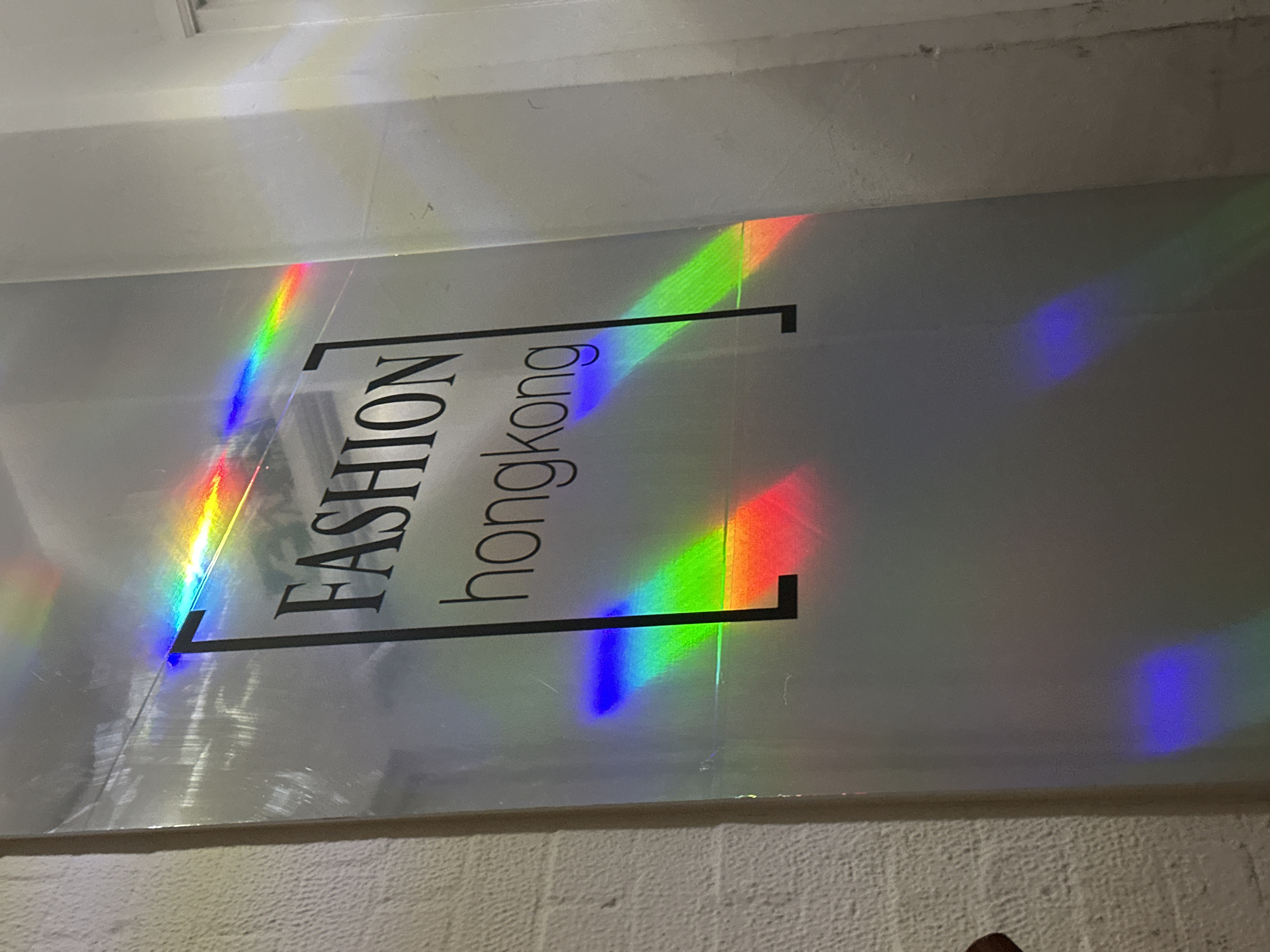 The image depicts a vertical sign displaying "FASHION hongkong" enclosed within a rectangular border. Rainbow light streaks overlay the sign and the surrounding wall, exhibiting the full spectrum of colors. The upper wall and lower brick wall appear textured, with the rainbow refractions seemingly caused by light passing through a prism or similar object.