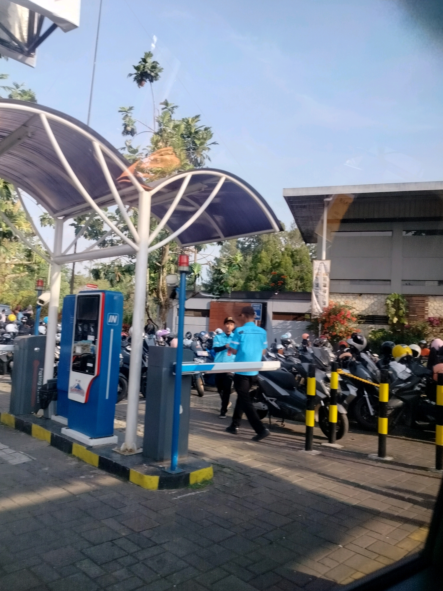 The image shows an outdoor parking area at a facility in Megamendung, Indonesia, likely a tourist attraction or public space. The scene features a covered parking shed with a blue and white ticket dispenser and a red and white barrier arm. Several motorcycles are parked in designated rows. Two individuals in blue shirts are standing near the barrier, and another person in a blue shirt is walking away from the ticket dispenser. The environment is characterized by paved ground, trees, and a building in the background. The sky is clear and blue, indicating a sunny day.