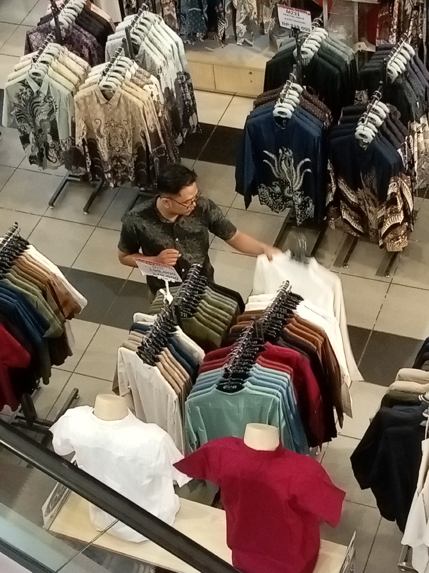 In Batam, Indonesia, a man in a dark, patterned short-sleeved shirt and glasses is standing in a retail store, interacting with clothing racks. He is holding up a white shirt. The store displays a variety of batik shirts on hangers and mannequins. The floor is tiled in a light color, and an escalator is visible in the foreground. The overall scene depicts a shopping environment focused on apparel.