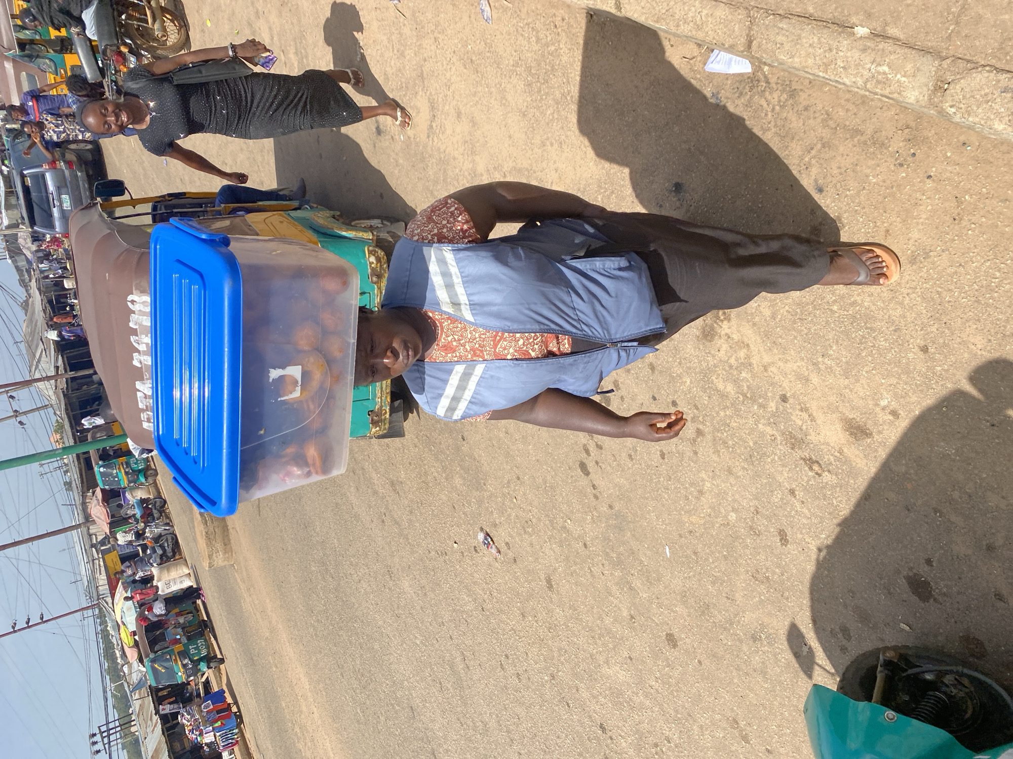 In a dusty outdoor marketplace in Bukuru, Nigeria, a woman is captured in a medium shot, walking towards the camera. She is carrying a large clear plastic tub filled with round, orange objects on her head. The tub is covered with a blue lid. The woman wears a blue vest over a patterned shirt and dark pants, with brown sandals on her feet. To her right, another woman in a black dress and white sandals walks away from the camera, smiling. In the background, several auto-rickshaws, motorcycles, and market stalls are visible, suggesting a busy commercial area. The ground is a mixture of dirt and asphalt, with scattered debris. A turquoise motorcycle is parked to the left of the frame. The scene is bathed in bright sunlight, casting long shadows.