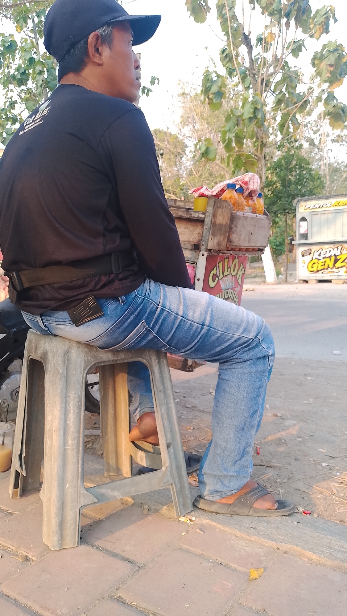 A man wearing a black cap, black long-sleeved shirt, and blue jeans sits on a grey plastic stool on the side of a road in Karanglangu, Indonesia. He is looking to his right, away from the camera. Behind him is a food cart with several bottles of orange liquid and a checkered cloth. Further in the background, there is a white and yellow food stall with the text "KEDAI GEN Z". The ground is paved with square bricks and dirt, with some scattered debris. Trees with green leaves are visible in the background under a bright sky. The lighting suggests it is daytime, likely late afternoon due to the long shadows.