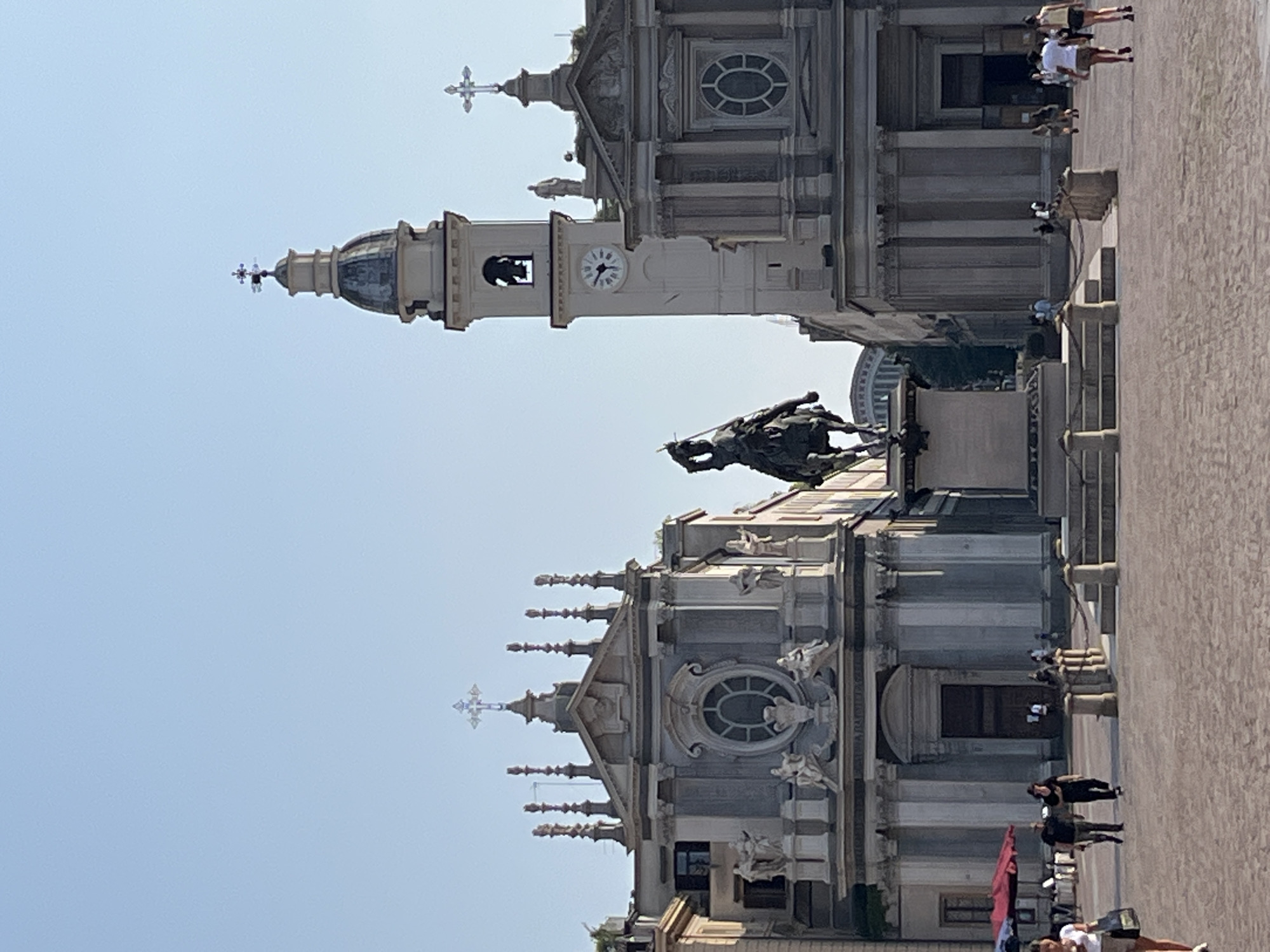 The image depicts Piazza San Carlo in Turin, Italy, on a clear day. The central focus is an equestrian statue positioned in the foreground, overlooking a wide, paved square. On either side of the statue are two Baroque churches, facing each other across the piazza. The church on the left features a prominent facade with intricate sculptures and a large oval window, topped with a cross. The church on the right is similar in architectural style, with columns, ornate details, and a cross on its roof. Between the two churches, a tall bell tower with a clock face rises into the sky, also adorned with a cross at its apex.
The square is populated with several individuals engaged in various activities. In the foreground, a woman in a white top and beige skirt walks from left to right, carrying a bag. To her left, two men are walking towards the camera; one wears a dark t-shirt and shorts, the other a black t-shirt and dark pants. Further back, near the entrance of the church on the left, an open red and white umbrella is visible, with tables and chairs set up nearby, suggesting a potential outdoor seating area. Other pedestrians are scattered across the piazza, some walking, others standing. The overall atmosphere is calm and leisurely, indicative of a public space being enjoyed on a sunny day. The architecture and layout of the piazza are consistent with the historical significance and grandeur of Turin's city center.