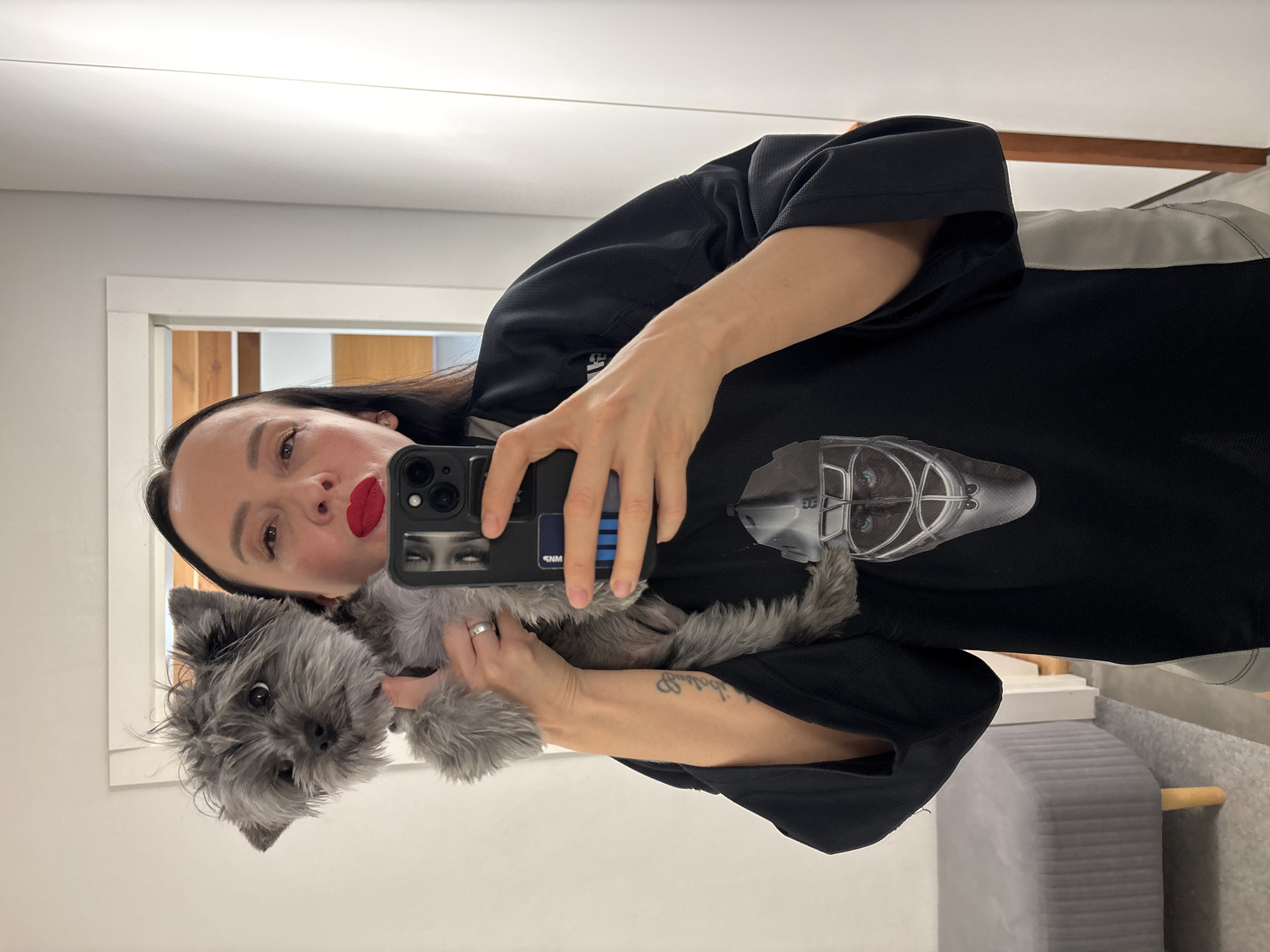 A woman with dark hair and red lipstick is taking a mirror selfie while holding a small, fluffy grey dog. She is wearing a black, short-sleeved top featuring a stylized, metallic-looking helmet or head graphic on the front, with faint letters like "BE" and "CG" visible within the design. She holds a black smartphone with her right hand, which has a phone case depicting eyes and the letters "SNM" along with blue stripes. A silver-colored ring is visible on her left ring finger, and a script tattoo is on her right forearm.
The small dog, with light grey fur, looks towards the camera with a neutral expression. The setting appears to be indoors, likely a room with bright, even lighting, suggesting daytime. The background shows a white wall and a white door frame, beyond which a wooden door or wall element is visible. To the right, a grey, ribbed upholstered furniture piece, possibly a bench or ottoman, can be seen on a light-colored carpeted floor. The image is taken in Chaikovsky, Russia.