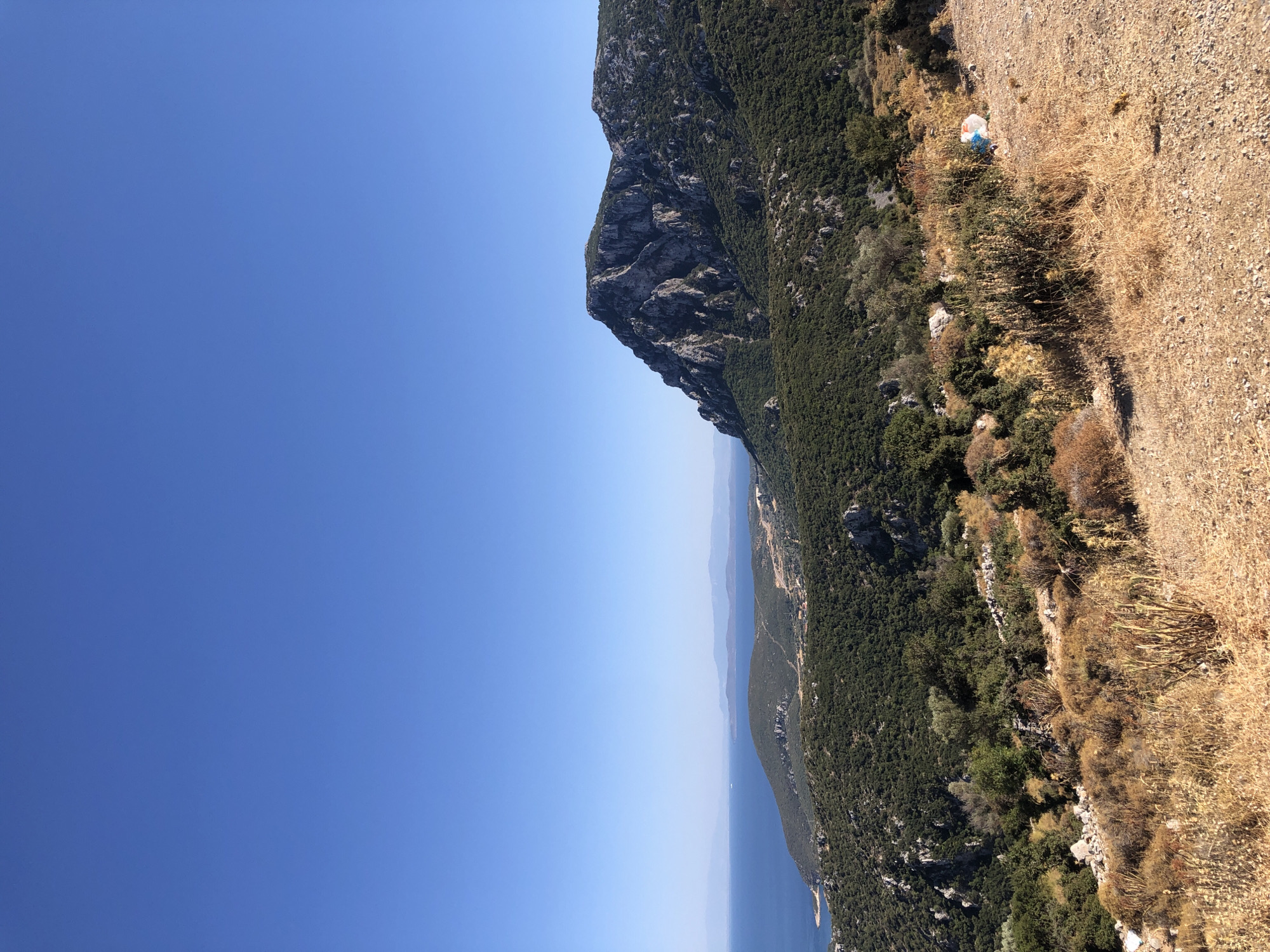 The image captures a panoramic view of a coastal mountainous landscape, likely in the vicinity of Karaburun, Turkey, given the geographical context. The foreground is dominated by dry, arid vegetation and a dirt path, characteristic of a Mediterranean climate during a dry season. In the mid-ground, rolling hills covered in dense green foliage, likely olive trees or similar Mediterranean shrubs, extend towards the sea. Scattered rock formations are visible amidst the greenery. A prominent, rugged mountain with exposed rock faces rises on the right side of the frame, its slopes also covered in vegetation. The background features the deep blue Aegean Sea meeting the horizon, with several islands and peninsulas visible in the distance, fading into a hazy, atmospheric perspective. The sky is a clear, vibrant blue. A piece of white and blue trash is visible on the right side of the path in the foreground.