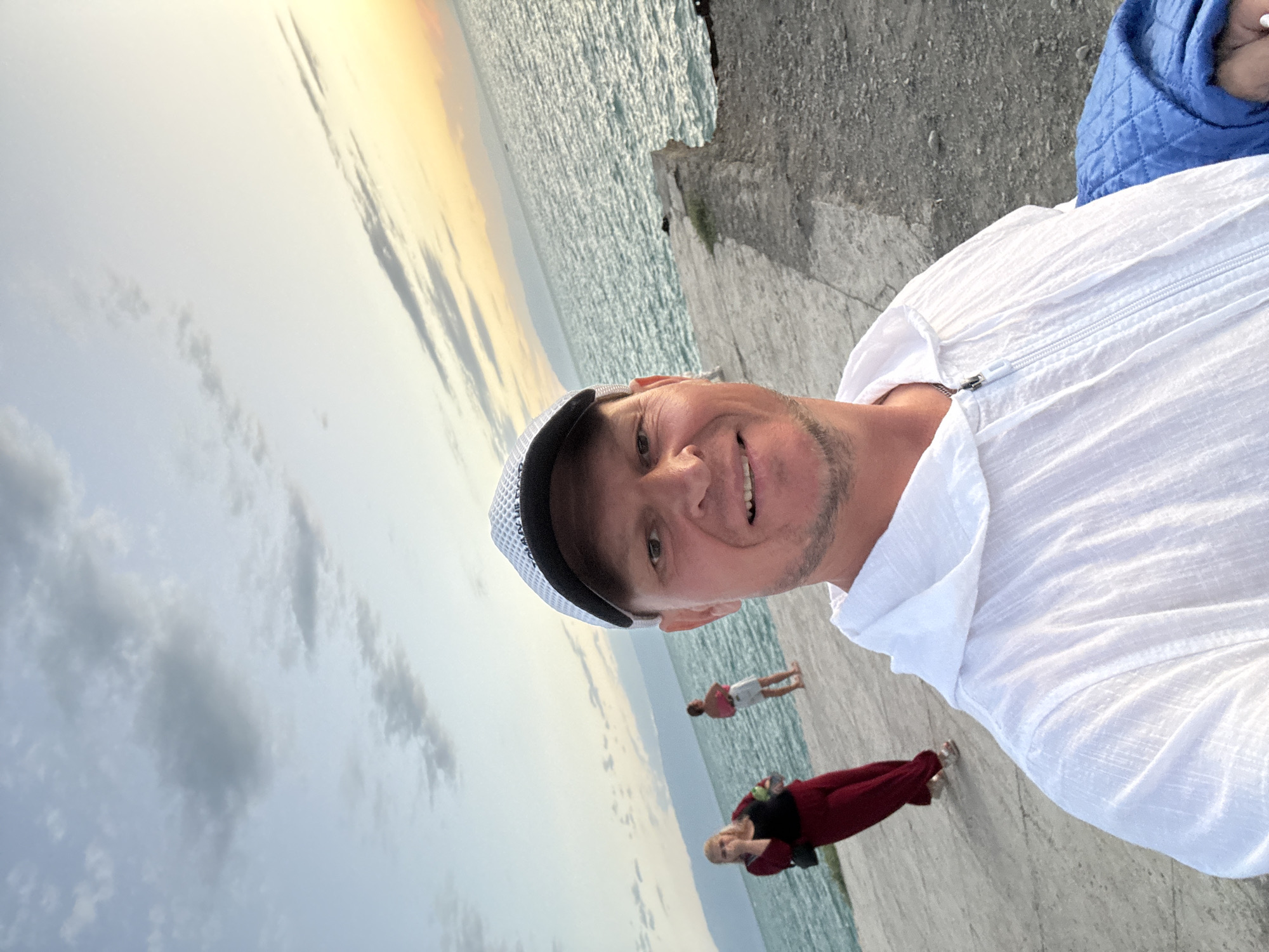 A man with fair skin, wearing a white hooded zippered jacket and a black and white baseball cap, is taking a selfie. He has short brown hair, stubble, and a light smile. Behind him, two women are visible on a concrete pier next to a body of water. The woman on the left is wearing a red outfit and the woman on the right is wearing a white skirt and pink top. The water appears calm, and mountains are visible in the distance. The sky is partly cloudy with a gradient of colors from light blue to yellow, indicating it may be sunset or sunrise. The location is identified as Zharbulak, Kazakhstan.