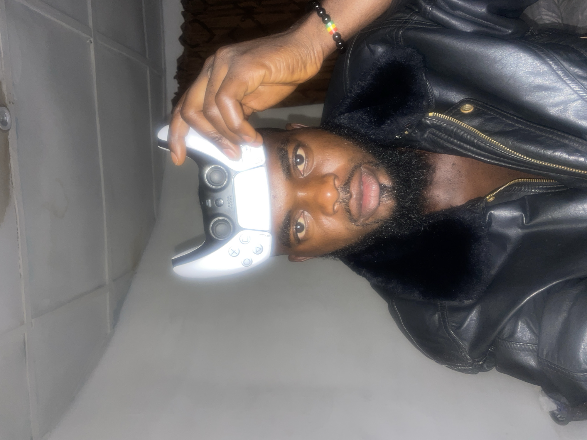 A dark-skinned man is positioned indoors, holding a white PlayStation 5 controller above his head. He is wearing a black leather jacket with a fur collar. He has a black beaded bracelet with red, yellow, and green beads on his wrist. The background consists of a white tiled wall on the left and a textured brown surface at the top. It is not possible to determine if this is a photo from Jos, Nigeria.
