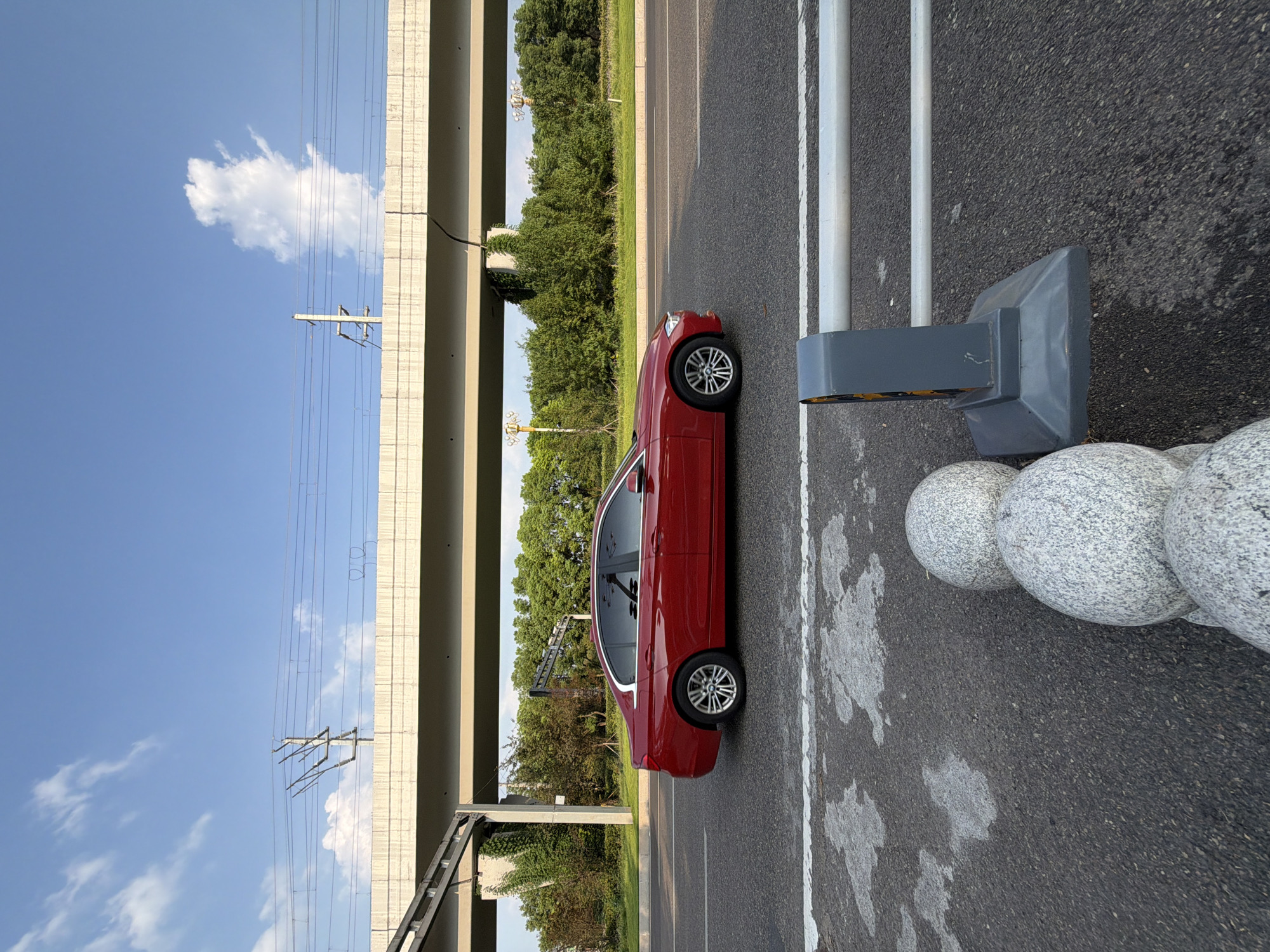 A red sedan drives on a road in Jiangjin, China, beneath an overpass with power lines. The sky is blue with scattered white clouds. Trees and vegetation line the road behind the overpass. In the foreground, a concrete barrier with two horizontal pipes is visible, along with three spherical concrete bollards.