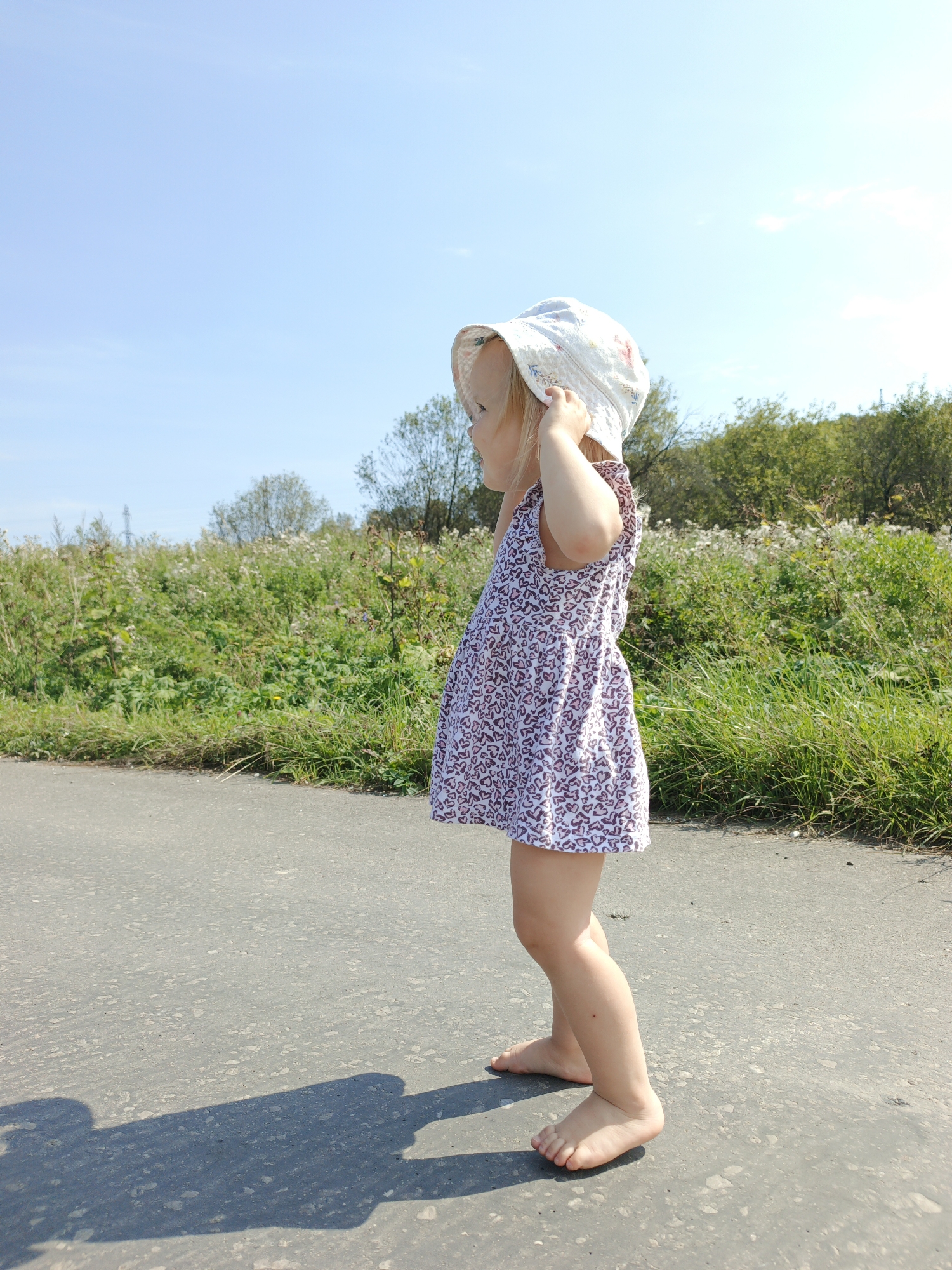 A young child wearing a white, floral-patterned sun hat and a short, sleeveless dress with a pink and white heart pattern stands on an asphalt road. The child is facing away from the camera, looking to the left. Their hands are raised to their head, adjusting the hat. The child is barefoot. The road stretches into the foreground and mid-ground, with a grassy area filled with wildflowers and trees bordering it on the right side. The sky is a clear blue. The scene appears to be in Urtiushka, Russia, given the context.