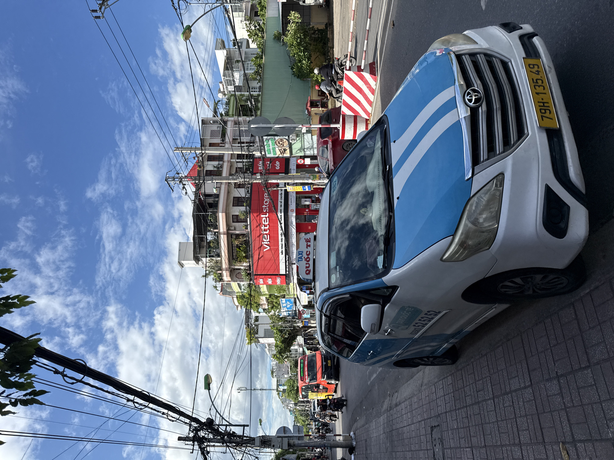 This image captures a street scene in Nha Trang, Vietnam, on a bright, partly cloudy day. In the foreground, a white and blue taxi, possibly a Toyota Innova model, is parked on the side of the road. The taxi displays the number "79H-135.49" on its yellow license plate. On the side of the taxi, the words "TAXI QUỐC TẾ" and a phone number "0258 52 52 52" are visible.
The street is paved with dark grey tiles on the sidewalk and asphalt for the road. A red and white striped barrier is situated in the middle of the road. A red minivan, also branded as a taxi, with "XA LỘ VIỆT NAM" written on it, is driving on the road alongside other vehicles and motorcycles.
The background reveals a bustling urban environment with multi-story buildings. A prominent red billboard for "Viettel Store" is visible, indicating a commercial area. Overhead, a complex network of power lines and streetlights crisscrosses the sky, with green foliage from trees peeking out from the top left. Several people are seen on motorcycles. The architecture suggests a tropical climate.