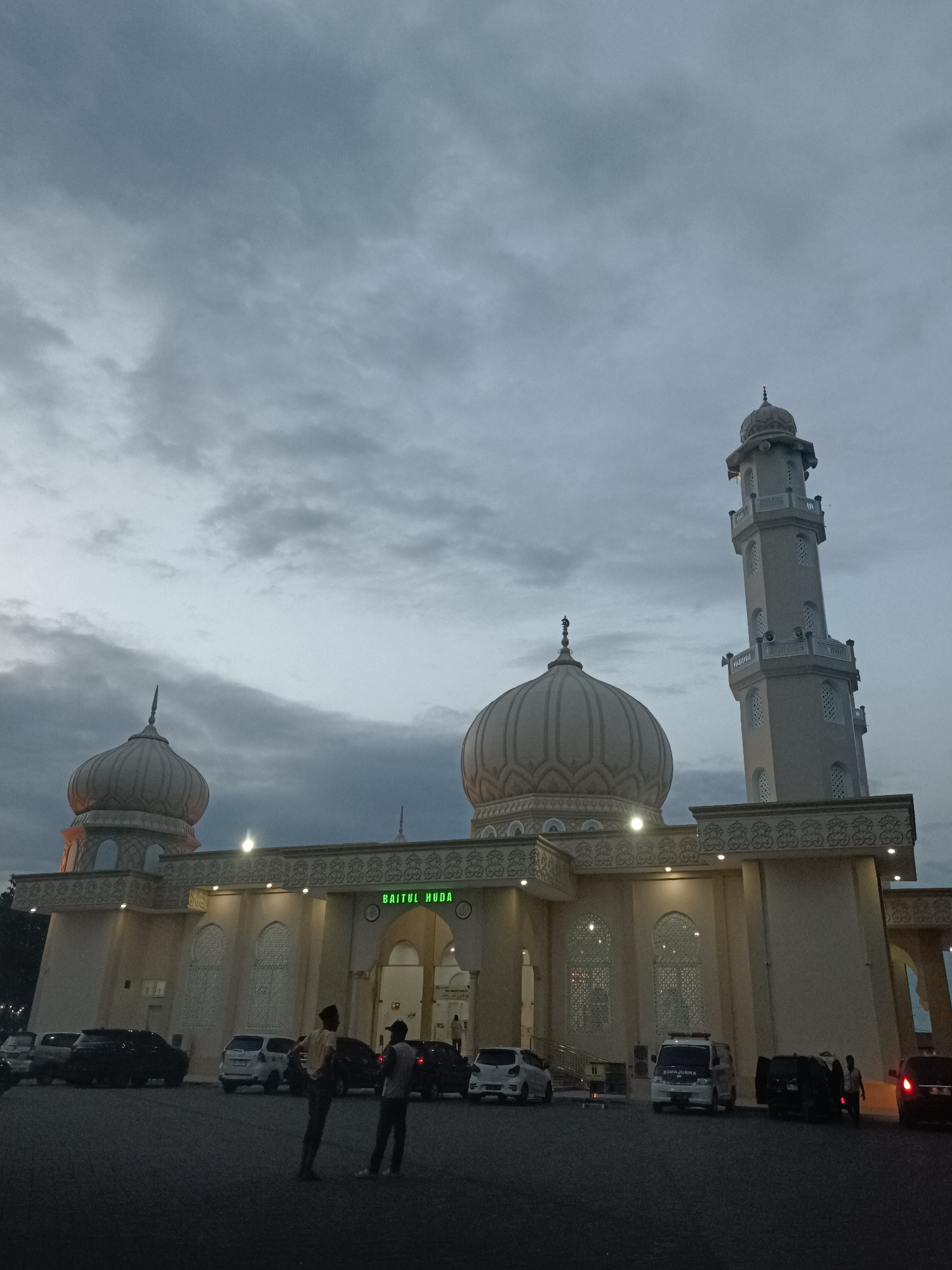 The image depicts the Baitul Huda mosque in Mee Tanjong Usi, Indonesia, at dusk. The sky is overcast with a muted gray tone. The mosque is a prominent, light-colored structure with a tall minaret and two large domes. Intricate decorative patterns adorn the building's facade, and lights are visible along the roofline, illuminating the architecture.
In the foreground, a dark paved area serves as a parking lot. Several vehicles are parked, including a white van that appears to be an ambulance, and other cars. Two individuals are standing in the parking lot, facing each other and appearing to be in conversation. Their presence suggests a quiet, possibly end-of-day scene at the mosque. The overall atmosphere is serene, with the fading light and the imposing presence of the religious building creating a sense of peace. The text "BAITUL HUDA" is visible above the entrance to the mosque.