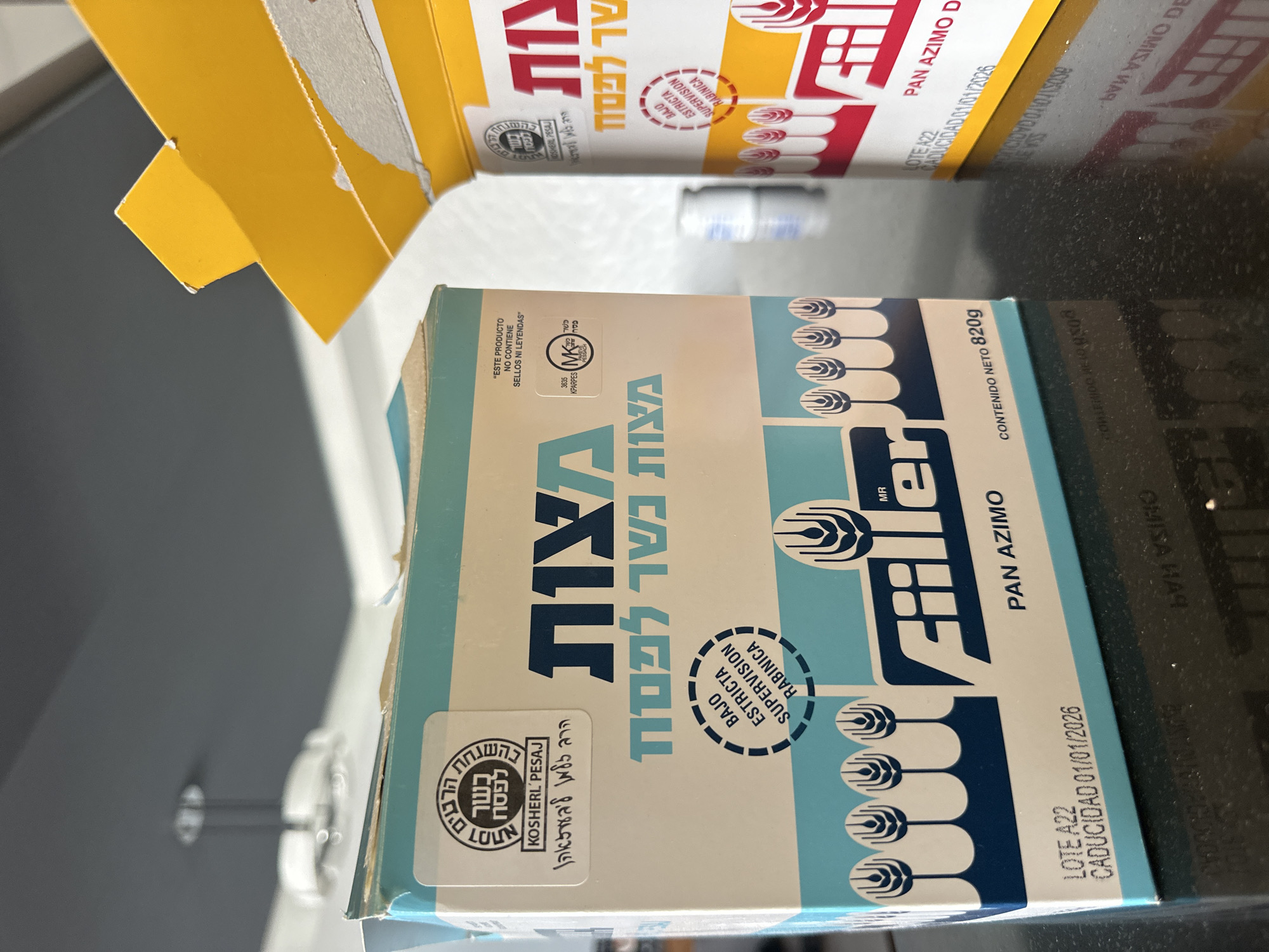 The image shows two boxes of "Filler" brand Pan Azimo matzah, stacked on a reflective black surface. The blue and white box is in the foreground, and the yellow and red box is partially visible in the background. The blue box has Hebrew writing and a Kosher certification seal. It lists the net content as 820g and has an expiration date of 01/01/2026. There is also Spanish text on the box stating the product contains no stamps or legends. The reflective surface shows a distorted reflection of the boxes. The scene appears to be indoors.