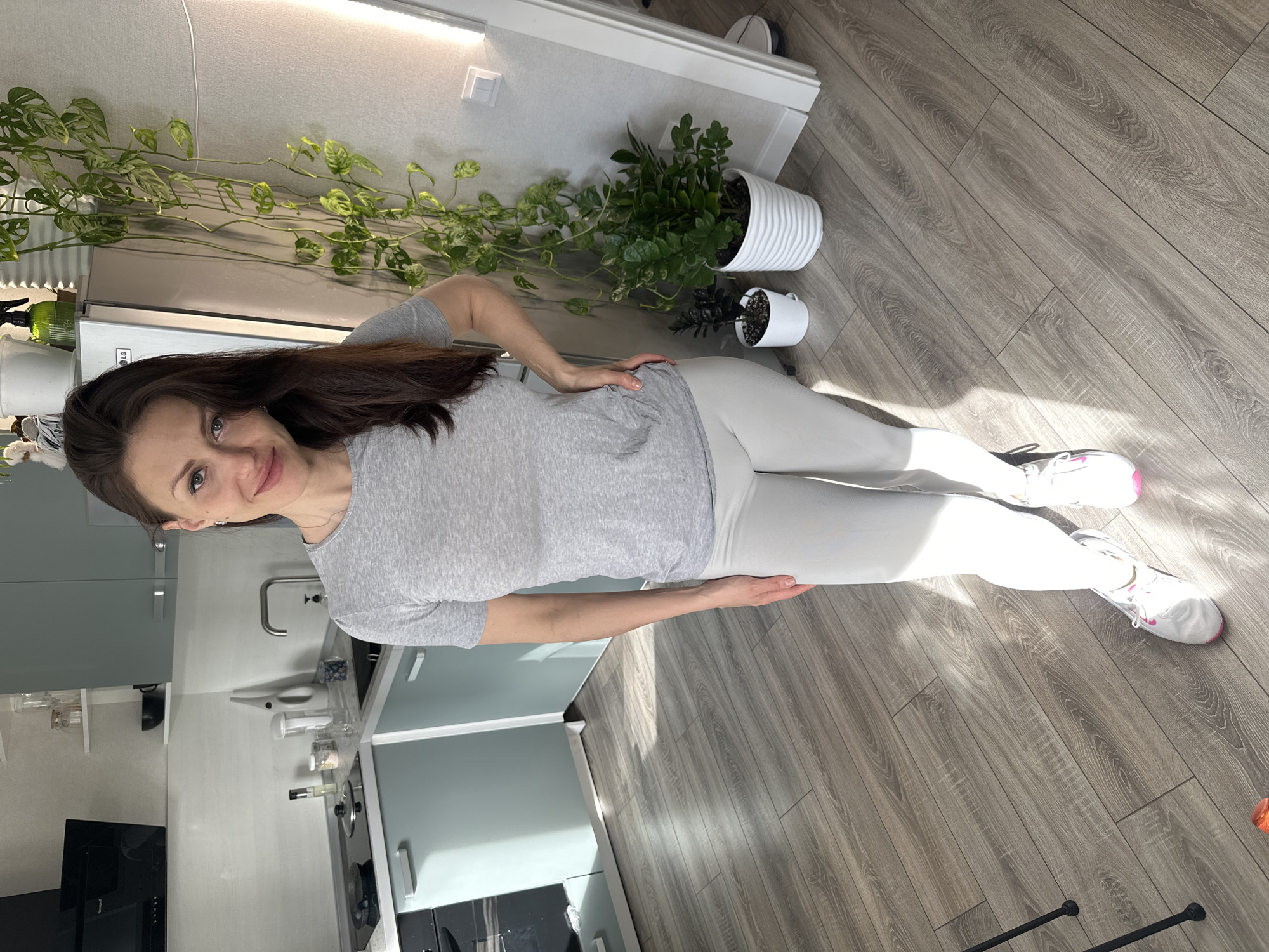 The photograph depicts a woman indoors in what appears to be an apartment. She is standing on a wood-patterned floor, wearing a gray t-shirt, light gray leggings, and white sneakers with pink accents. She is facing the camera with a slight smile. The woman's hair is dark brown and shoulder-length.
Behind her is a kitchen area with light blue cabinets, a stainless steel sink, and a black cooktop. On the wall above the cabinets, there is a plant with trailing vines. To the right of the plant are other potted plants. A white baseboard runs along the wall.
The location is identified as Murino, Russia.
