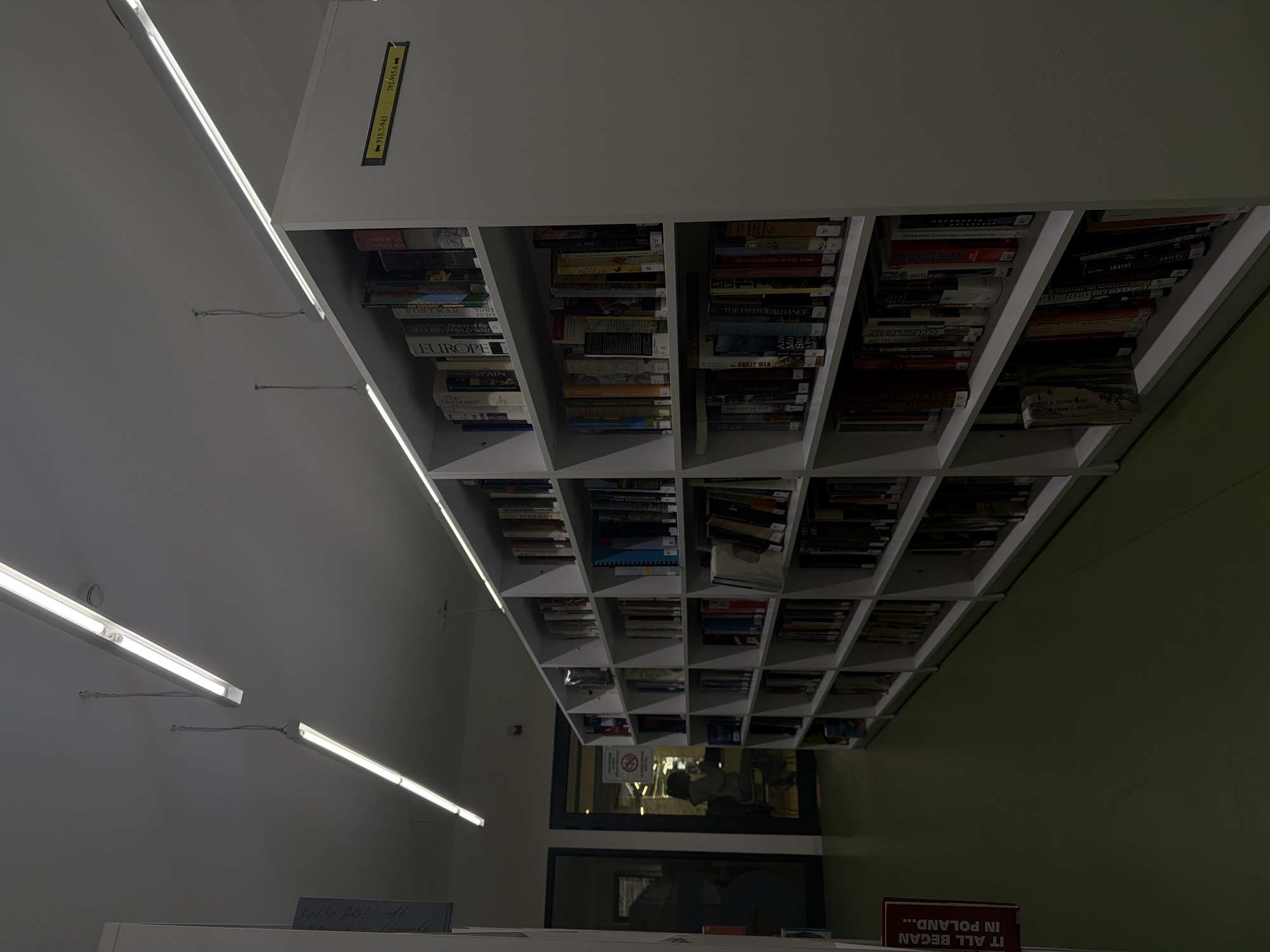 The image shows a library with ceiling-high bookshelves filled with books in an indoor setting. The location is unknown, but there is no evidence it is located in Yerevan, Armenia. The shelves are white and divided into cubicles, each containing books. Fluorescent lights illuminate the room. A window reflects a glimpse of people and the outside, and some signs are visible. A sign "IT ALL BEGAN IN POLAND" is visible on the shelf below. Some of the books are labeled "WAR" and "EUROPE."