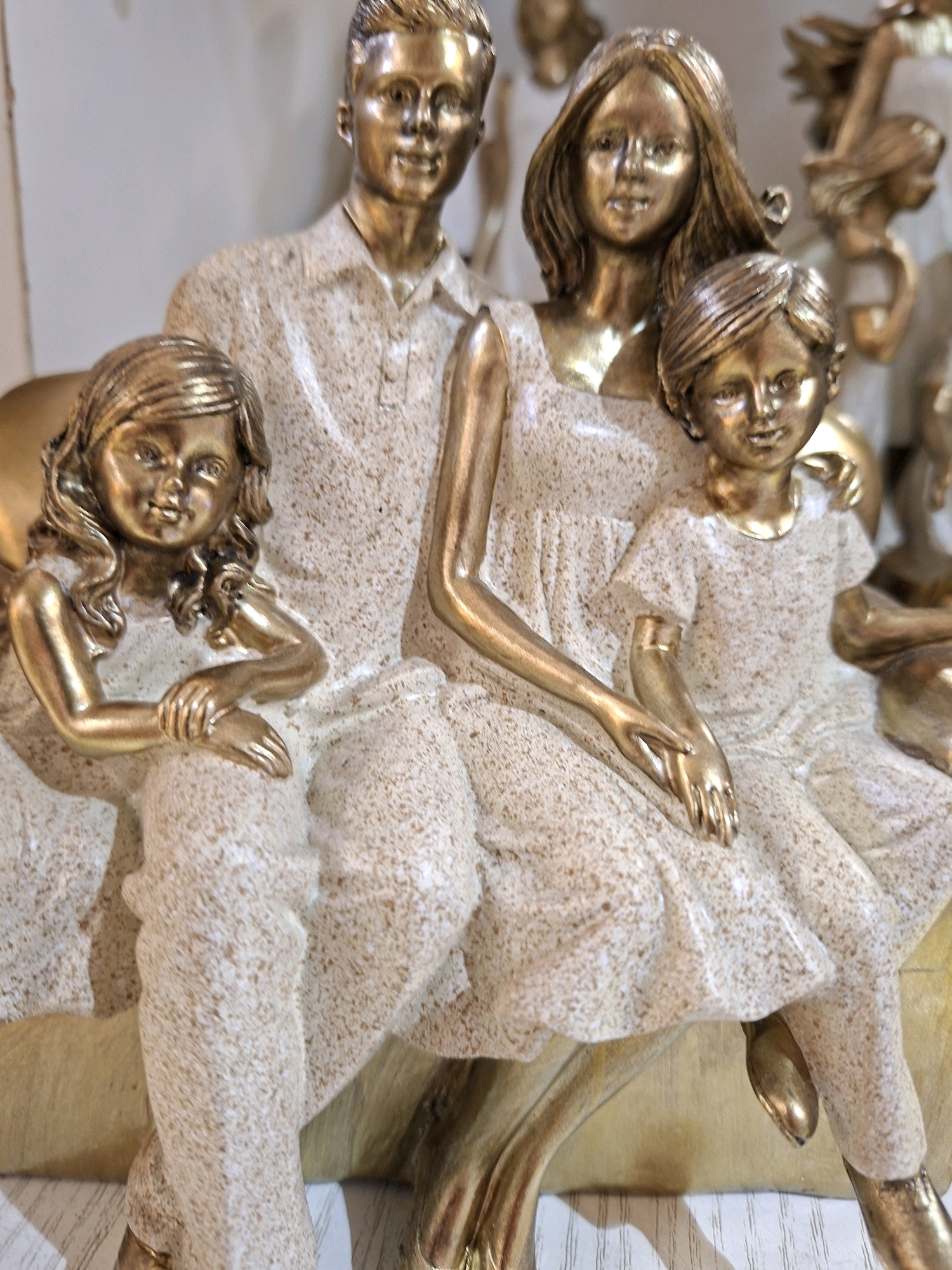 This is a close-up, low-angle studio photograph of a gold-colored resin sculpture depicting a family. The sculpture features a father, mother, and two children, all with a distressed, speckled gold finish. The father is standing, wearing a collared shirt. The mother is seated behind him, with a young girl in her lap. Another boy is seated in front of the parents, with his arm around the mother. The sculpture is resting on a base that appears to be part of the same material, with gold accents on the edges. The background is a plain, light-colored surface, possibly a wall or backdrop. The lighting is bright, creating strong highlights and shadows on the sculpture, emphasizing its metallic sheen and textured surface. There is no indication of the city of Cologne, Germany, in the image.
