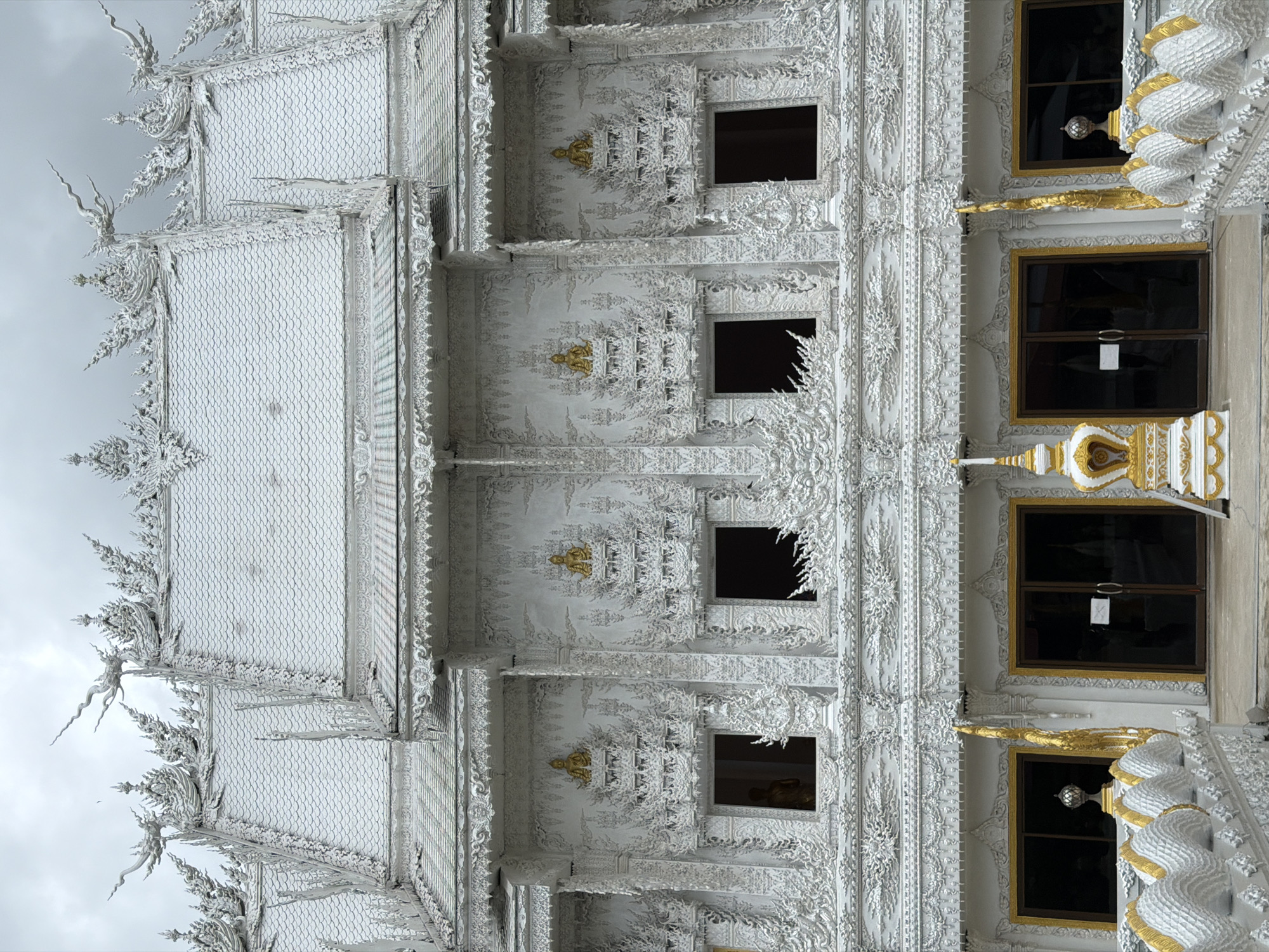 The image captures an upward view of a white, highly ornamented structure with golden accents. Four black framed windows are visible on the right side. The structure appears to be a building, possibly a temple. The architectural style features intricate carvings and sculptures throughout its exterior. The location is likely in Thep Krasatti, Thailand, as indicated in the prompt. The sky is overcast, suggesting daylight.