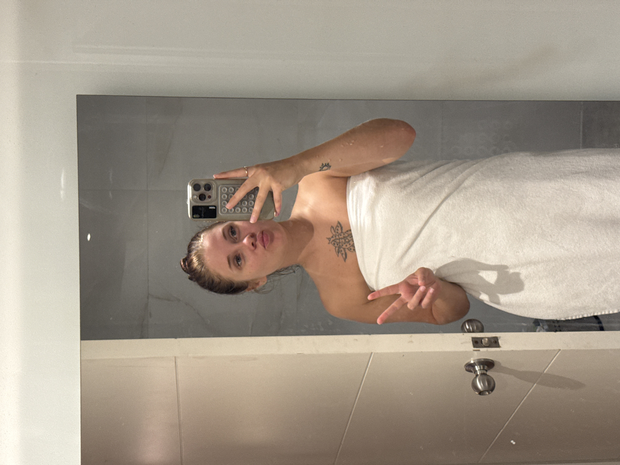 A woman is taking a mirror selfie in a bathroom. She is wearing a white towel wrapped around her body and has her hair pulled up in a bun. She is making a peace sign with her left hand and pouting her lips. She has a tattoo on her chest and a smaller tattoo on her right arm. The bathroom has light gray tiled walls and a white door with a silver doorknob. The image is taken from the chest up.