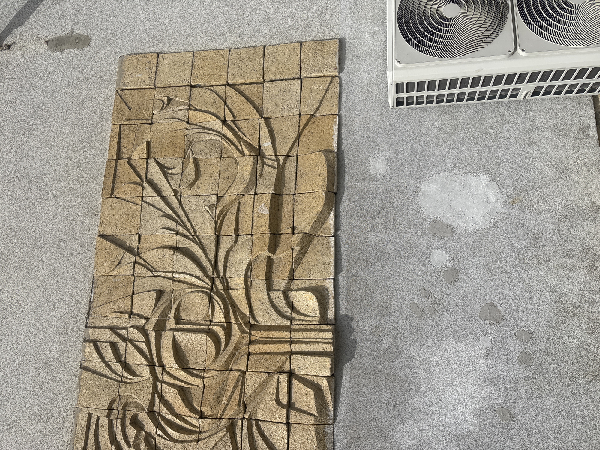 A stone wall mural is affixed to the side of a building in Troyan, Bulgaria. The mural is composed of square beige tiles, each carved with a stylized design of plants or vegetation. The mural is lit with sunlight, causing shadows to highlight the carved design. To the right of the mural is the building's concrete exterior, marked by a few patched spots. Above and slightly to the right of the mural is a modern air conditioning unit with two fans visible.