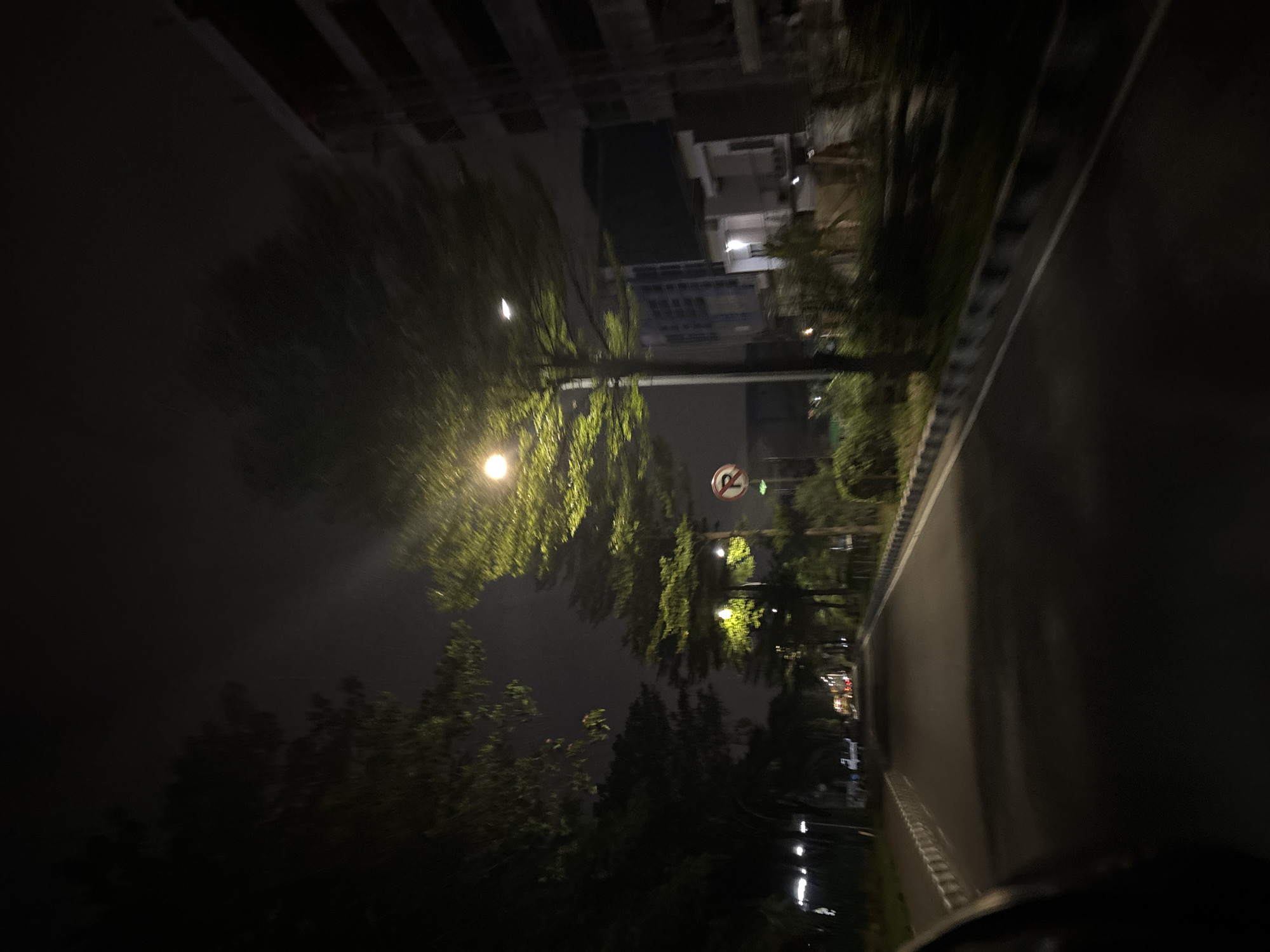 The image depicts a street in North Jakarta, Indonesia at night. A street lamp illuminates trees lining the road, casting light on their green foliage. A no-parking sign is visible next to the street lamp. Buildings stand in the background under a dark, cloudy sky. The road is wet, and street lights are reflected on the road surface.