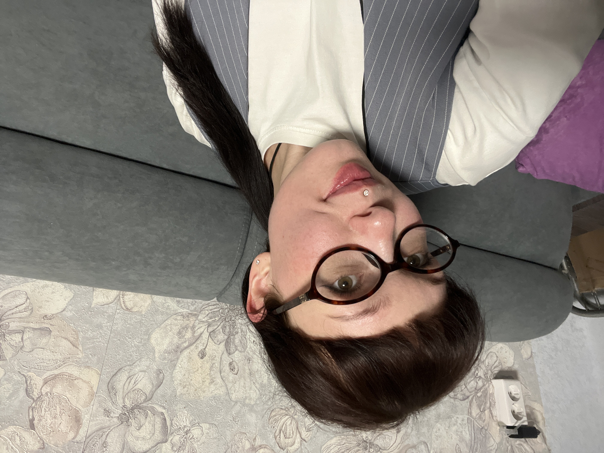 The image depicts an adult female indoors in Chaikovsky, Russia. The subject has dark brown hair styled with bangs and a ponytail, and wears oval tortoise-shell eyeglasses. A small, clear gemstone labret piercing is present on her lower lip, and a silver stud earring is visible in her left ear. She is wearing a white crew-neck t-shirt beneath a grey pinstriped vest, and exhibits a slight smile while looking directly at the camera. The background consists of a light-colored wall covered in wallpaper featuring a floral pattern. To the subject's left, a portion of a purple cushion is visible. A grey upholstered sofa runs horizontally behind the subject. In the upper left background, a white double electrical wall socket is mounted on the wall, with a black adapter plugged into the upper outlet and cables extending from both outlets.