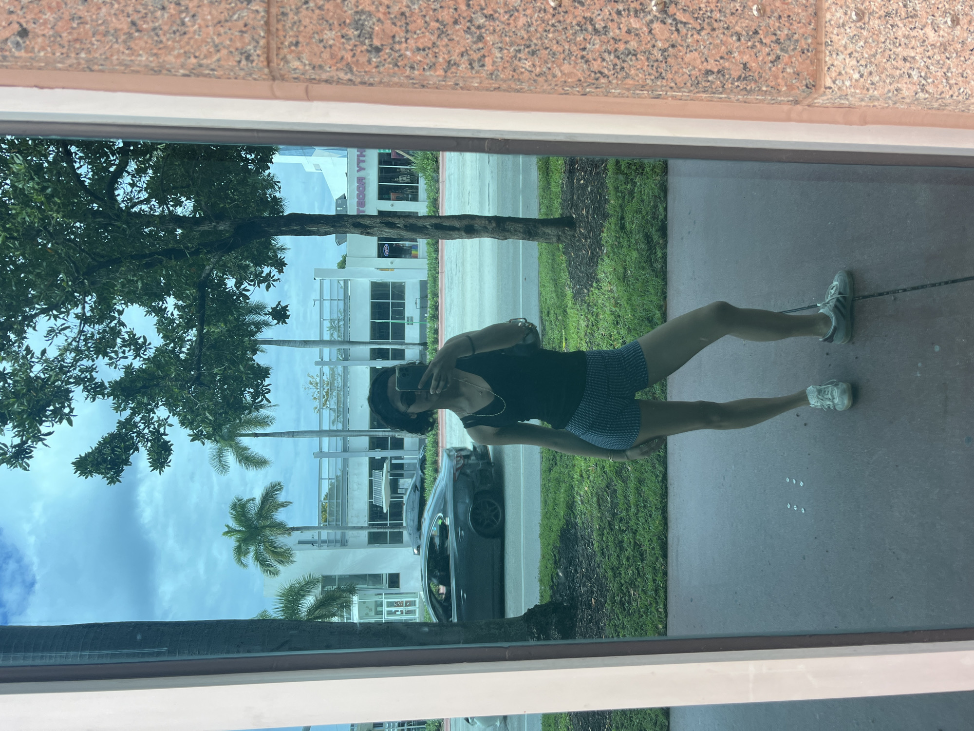 A woman in a black tank top and blue patterned shorts is taking a selfie in a reflective window. The reflection shows a street scene. In the background, there are buildings, palm trees, a road with a dark-colored sedan on it, and grassy areas on either side of the road. The architectural style and vegetation are consistent with Miami Beach, Florida, United States.