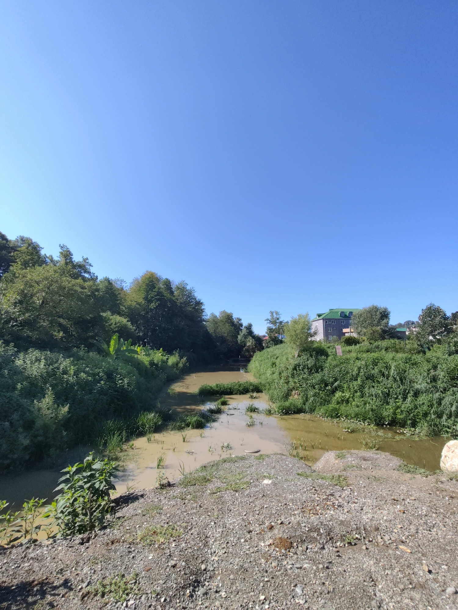 The image depicts a natural landscape with a brown, muddy river or stream flowing through lush green vegetation under a clear blue sky. On the right bank of the water, a modern, multi-story building with a green roof is visible, surrounded by trees and dense foliage. The foreground consists of a gravelly embankment. The scene appears to be located in or near the city of Oryol-Izumrud, Russia, though specific details of human activity are absent.