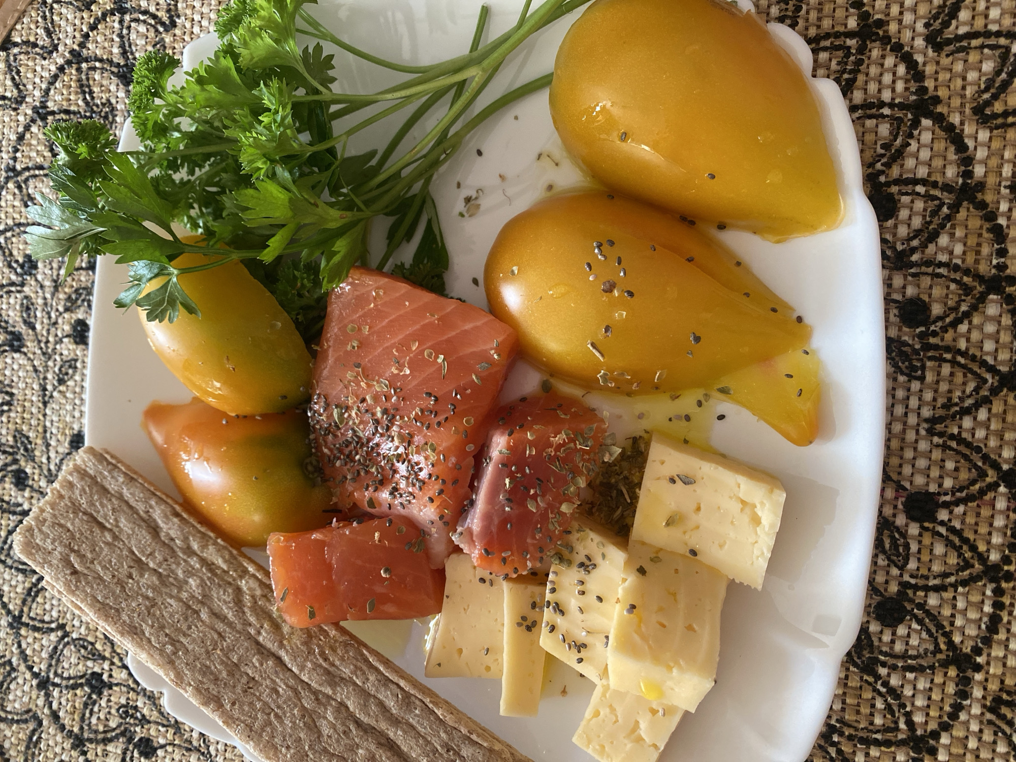 A close-up overhead shot shows a white plate on a patterned placemat. On the plate are several food items: three pieces of raw salmon seasoned with herbs and chia seeds, several cubes of yellow cheese, two whole yellow tomatoes drizzled with oil and sprinkled with seeds, a sprig of parsley, and a rectangular crispbread on the upper left. The scene appears to be indoors.