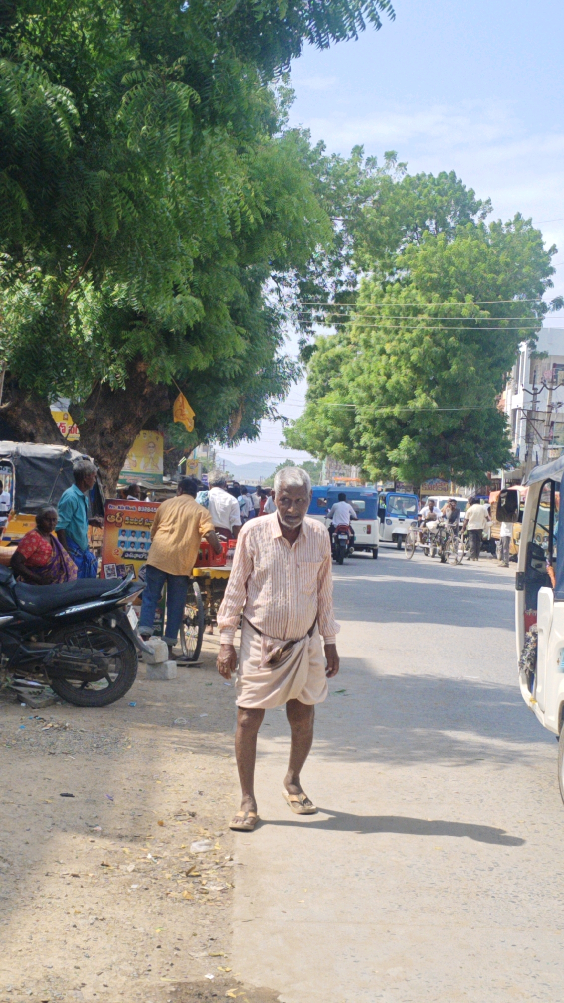 The image depicts a street scene in Virat Nagar, India. In the foreground, an elderly man with gray hair and a beard walks towards the viewer. He is wearing a striped, light-colored shirt and a light-colored dhoti wrapped around his waist. He is also wearing sandals. Behind him, to his left, a motor scooter is parked on the side of the road. Further back on the left, a woman in a red and blue sari is seated, and a man in a blue shirt and white pants stands next to her. A yellow and orange cart is positioned behind them, with a person wearing an orange shirt loading items from it. In the background, several vehicles, including auto-rickshaws and motorcycles, are on the road. The street is lined with trees, and buildings can be seen in the distance. The sky is clear and blue.