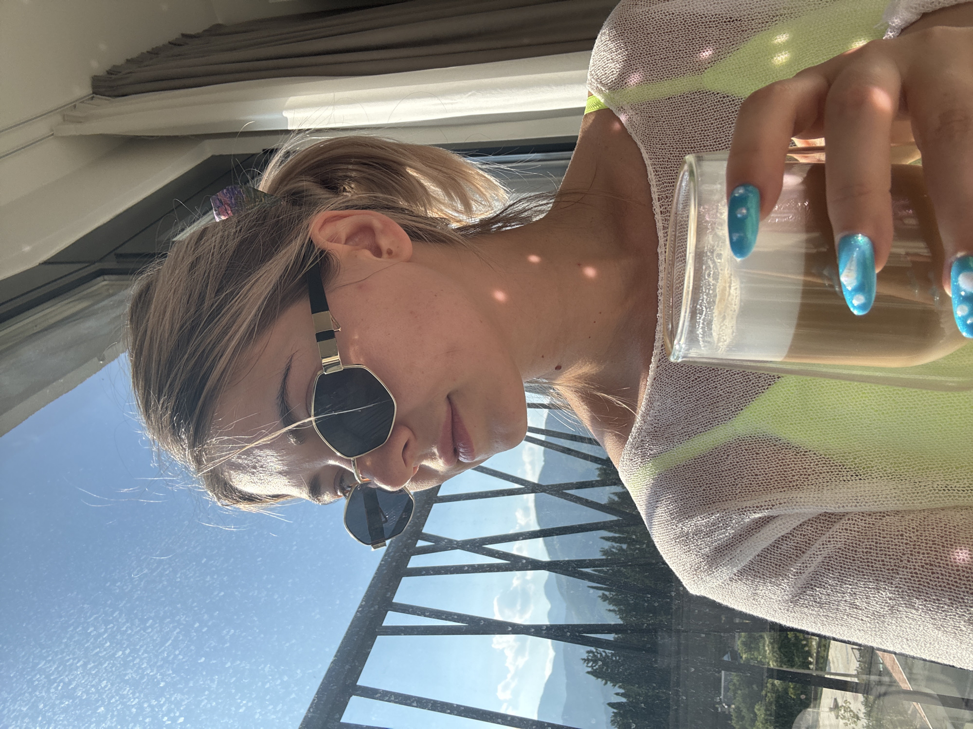 A woman with blonde hair pulled into a ponytail, wearing sunglasses and a sheer white top over a neon green garment, holds a glass of coffee in front of a window with a view of mountains and trees. Sunlight creates dappled shadows on her skin. The scene appears to be indoors, possibly a hotel room, with curtains visible in the background.