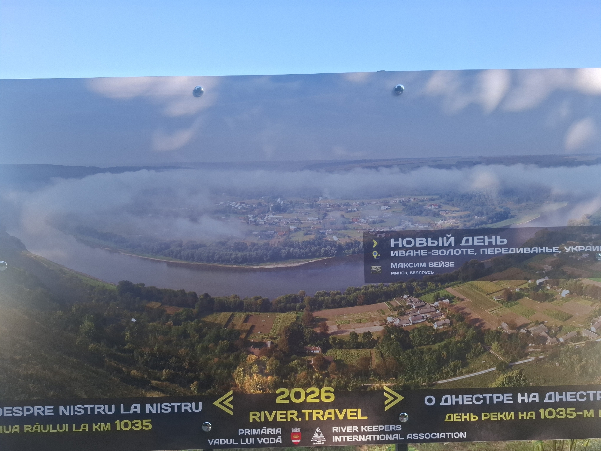 The image is a close-up, horizontal shot of a sign with a scenic aerial photograph of a river valley and a village. The sign has text in Russian and Romanian. At the top of the sign, some text in Russian reads "НОВЫЙ ДЕНЬ", "ИВАНЕ-ЗОЛОТЕ, ПЕРЕДИВАНЬЕ, УКРАИЯ", "МАКСИМ ВЕЙЗЕ", and "МИНСК, БЕЛАРУСЬ". Below this, in the center of the sign, is the year "2026" followed by "RIVER.TRAVEL". To the left of this, in Romanian, is "DESPRE NISTRU LA NISTRU" and "JIUA RAULUI LA KM 1035". To the right, in Russian, is "О ДНЕСТРЕ НА ДНЕСТР" and "ДЕНЬ РЕКИ НА 1035-М". At the bottom, in Romanian, is "PRIMARIA VADUL LUI VODA" and "RIVER KEEPERS INTERNATIONAL ASSOCIATION". The aerial photograph depicts a meandering river cutting through a lush green valley with a village nestled on its banks. The sky above is a pale blue with wispy clouds, and the sun is casting a soft light on the landscape. The sign itself appears to be made of metal and is mounted outdoors. There are visible metal bolts holding the sign in place.